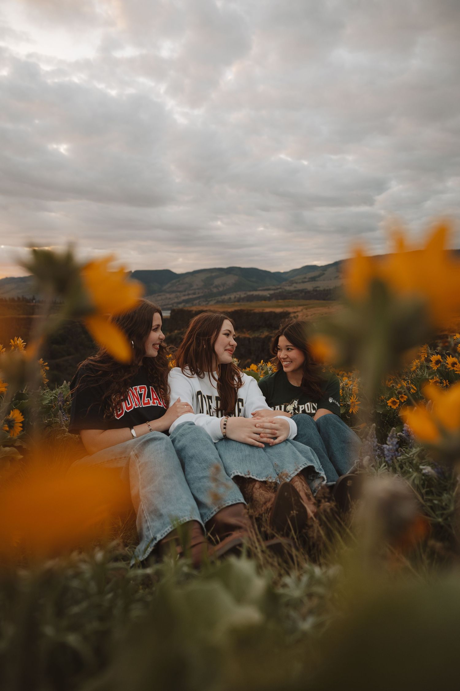 Three girls laughing together in a sunflower field with mountains and cloudy sky backdrop.