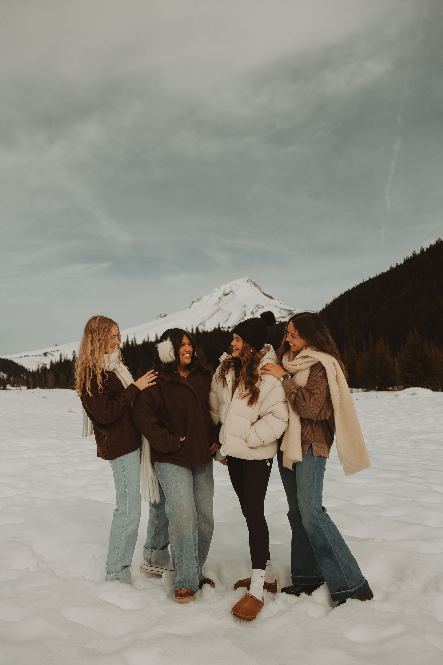 Five women in winter coats laugh together on a snowy field with a snow-capped mountain backdrop.