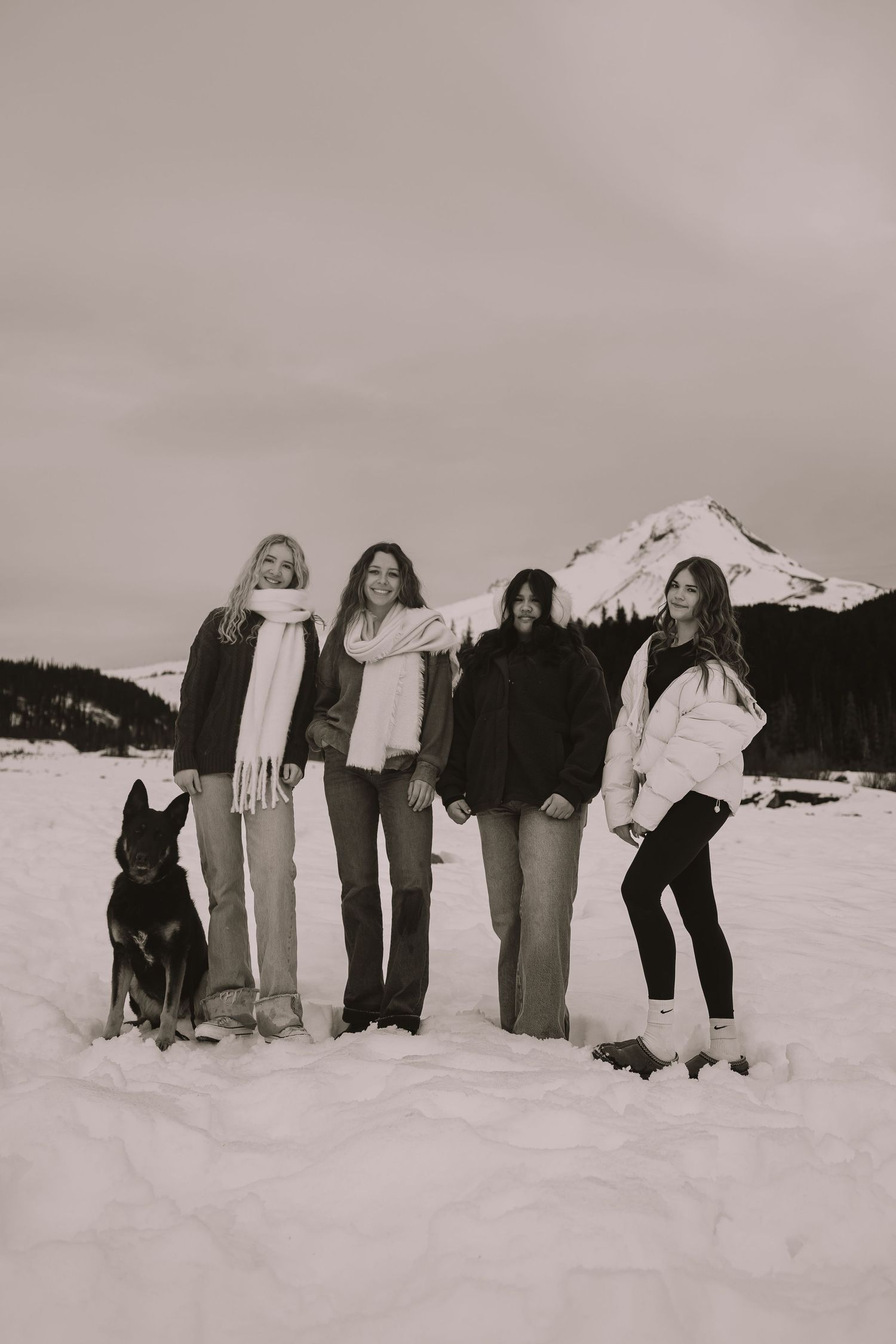 Four women and a dog pose together in black and white photo with snowy mountain backdrop.