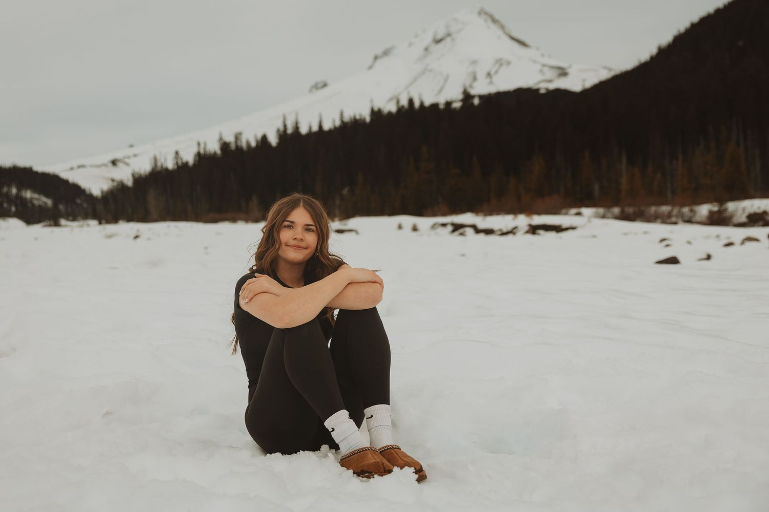 Young woman sits cross-legged on snow in mountain valley with evergreen forest and snowy peak behind her.
