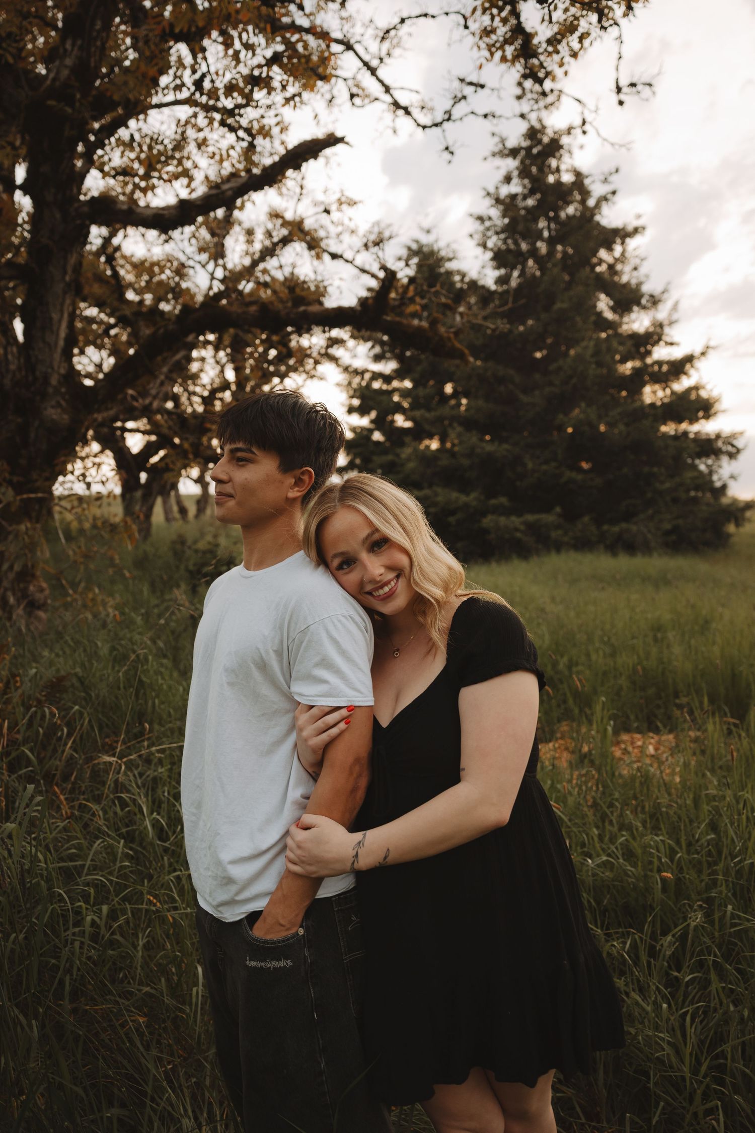 Couple embracing in a lush green field at golden hour, woman in black dress smiling at camera, man in white tee.