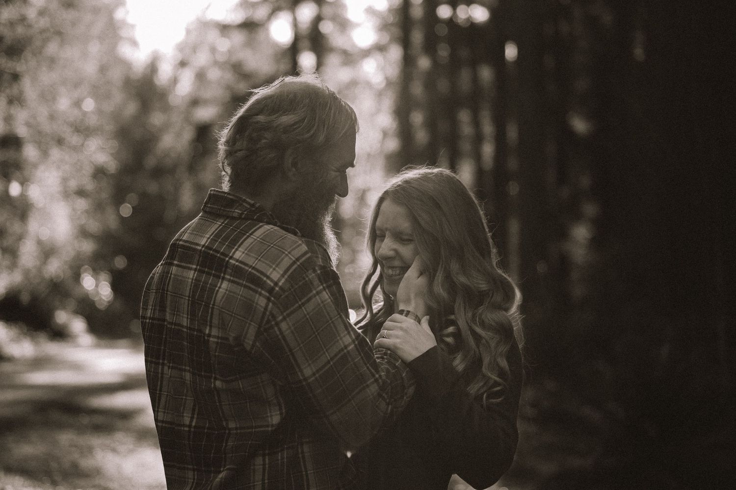 A couple in plaid clothing embraces in a romantic outdoor forest setting captured in black and white photography.