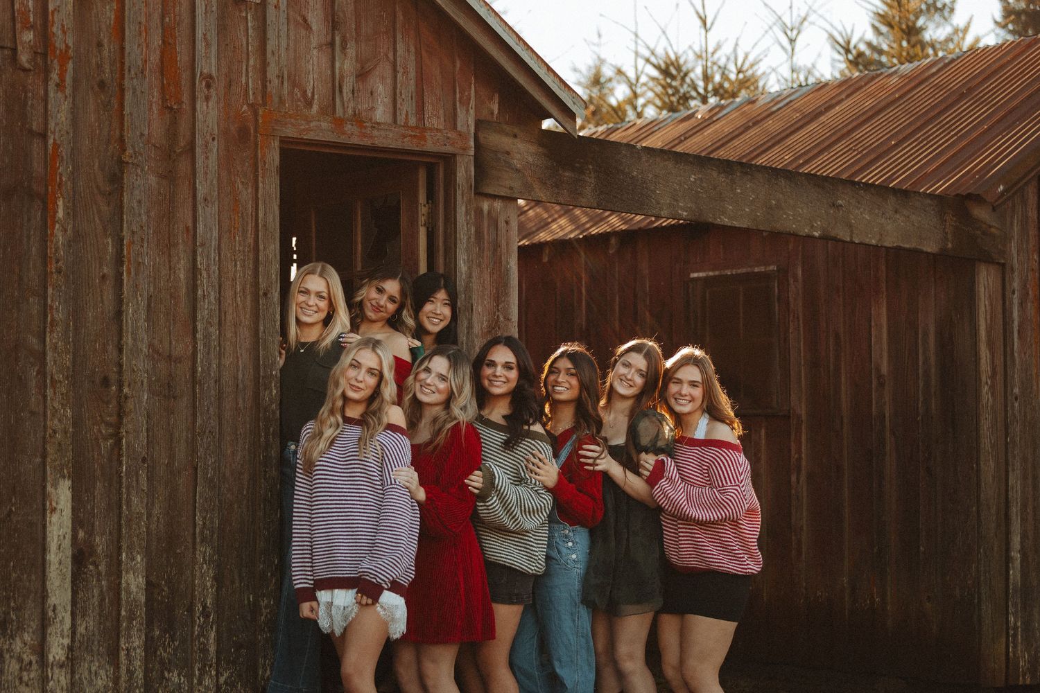 A group of friends in casual sweaters and shorts pose together in front of a rustic wooden barn at sunset.