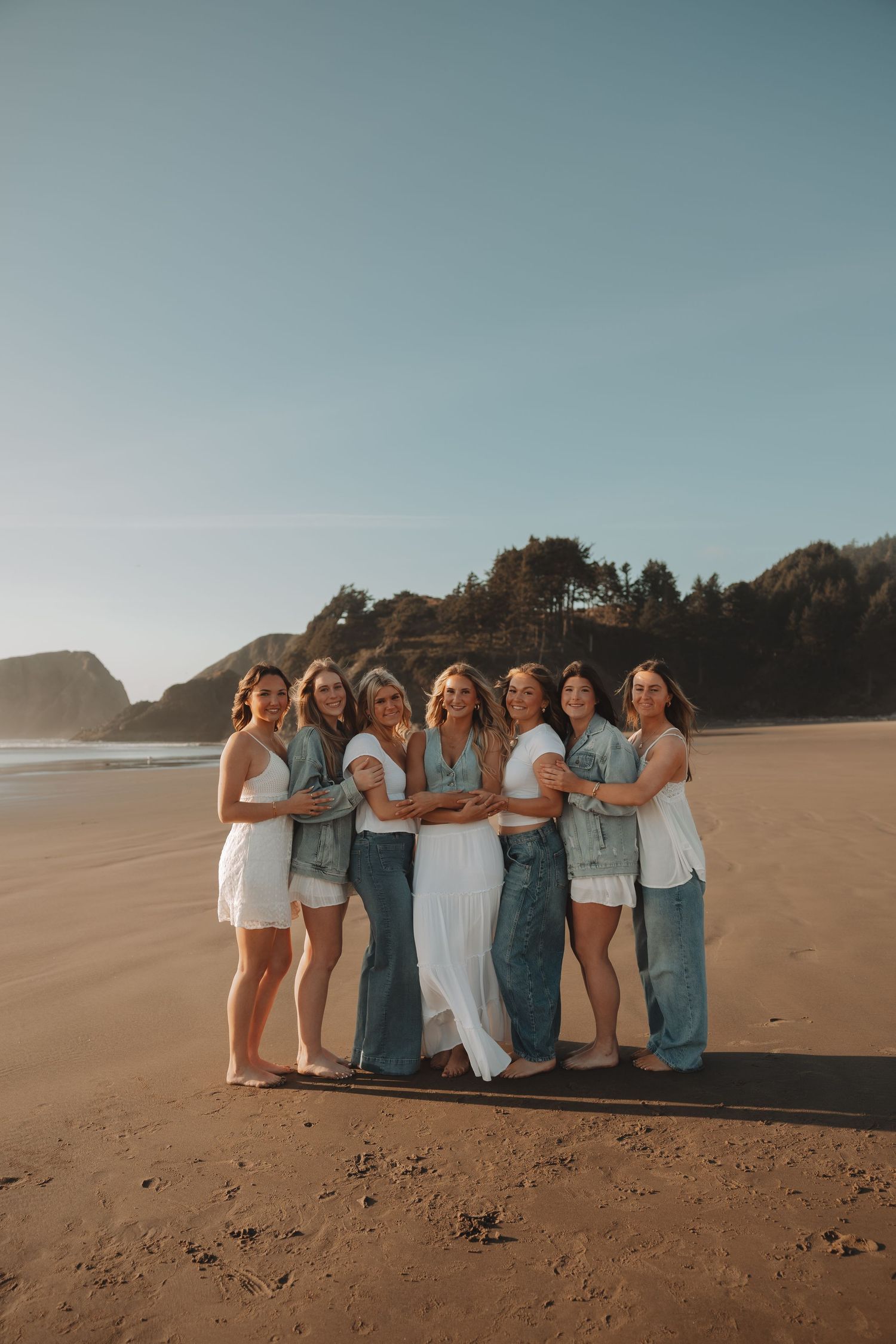 Group of eight women posing together on a sandy beach at golden hour, wearing white and denim outfits.