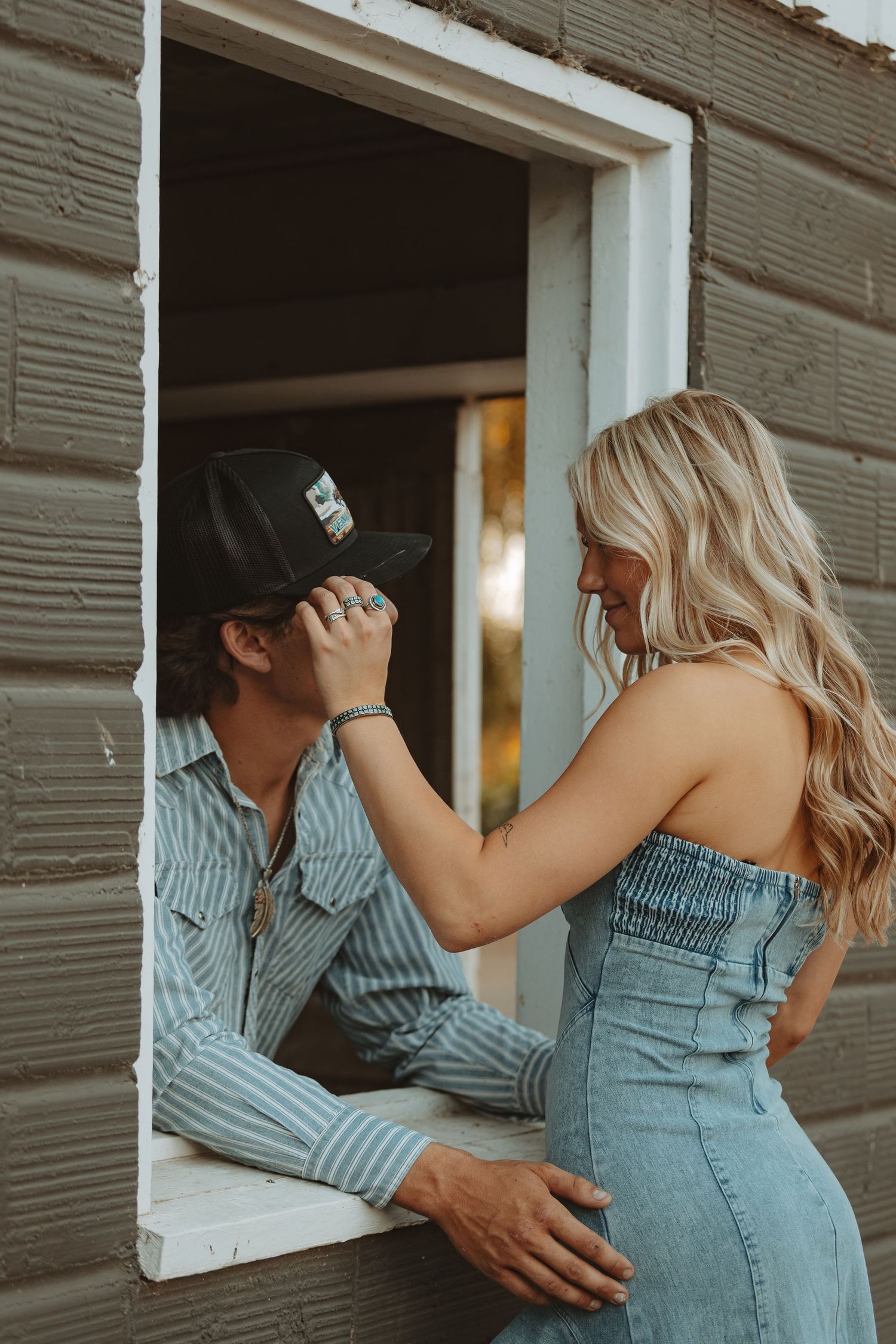 Blonde woman adjusting her partners cowboy hat through a rustic barn window at golden hour.