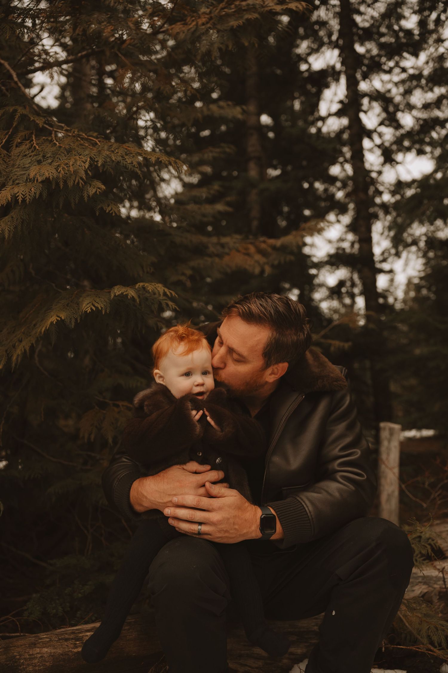 A couple shares an intimate moment in a moody forest setting with warm autumn tones and evergreen trees in the background.