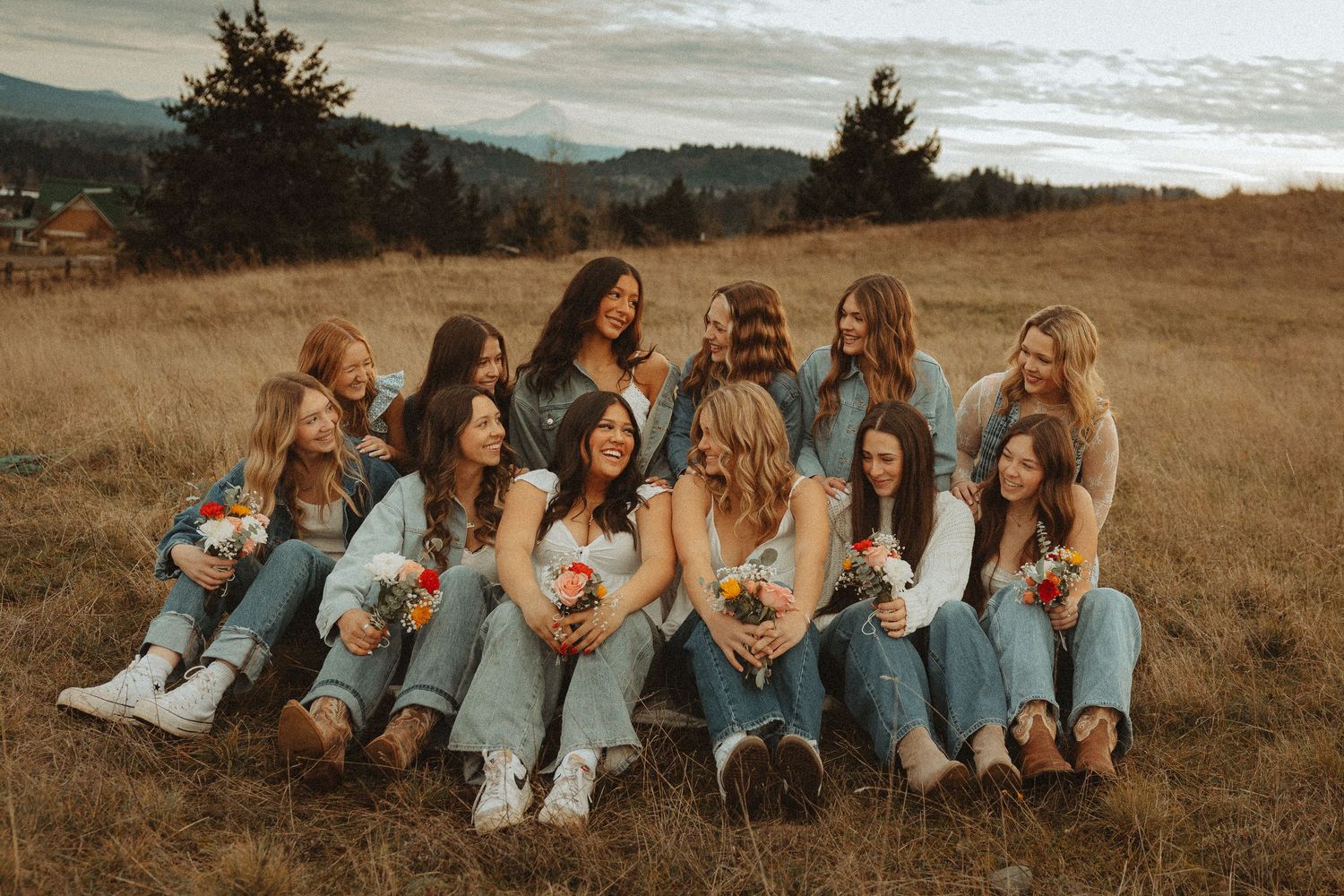 Friends in casual denim outfits sit together on a grassy hillside during golden hour.