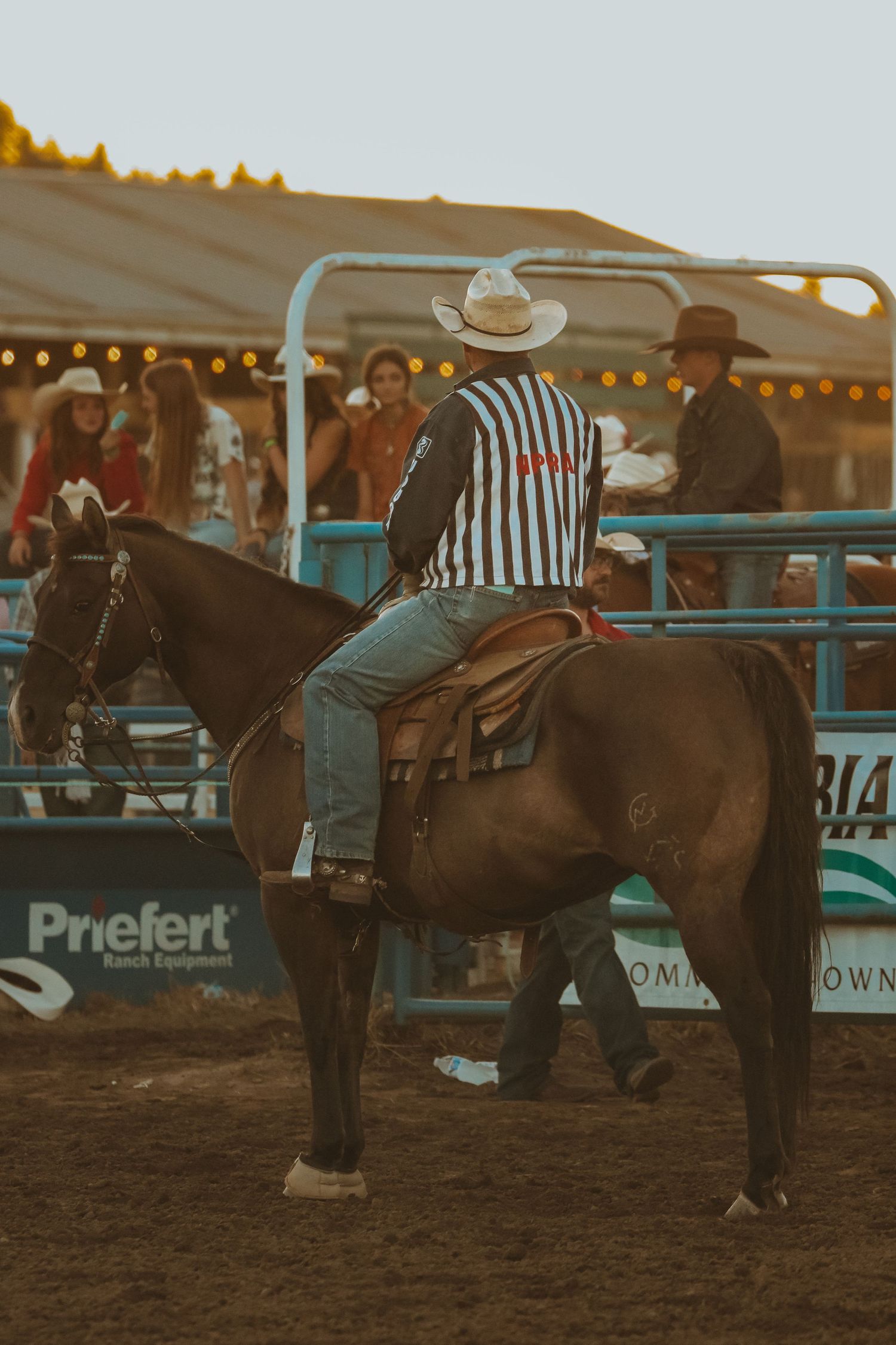 Rodeo officials and horses gather near the arena gates during sunset with equipment visible.