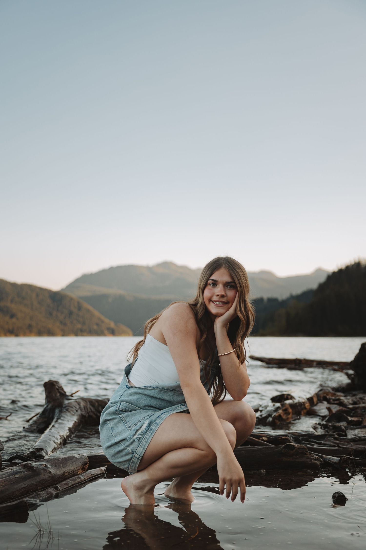Young woman in denim skirt sitting on driftwood by a mountain lake at dusk.