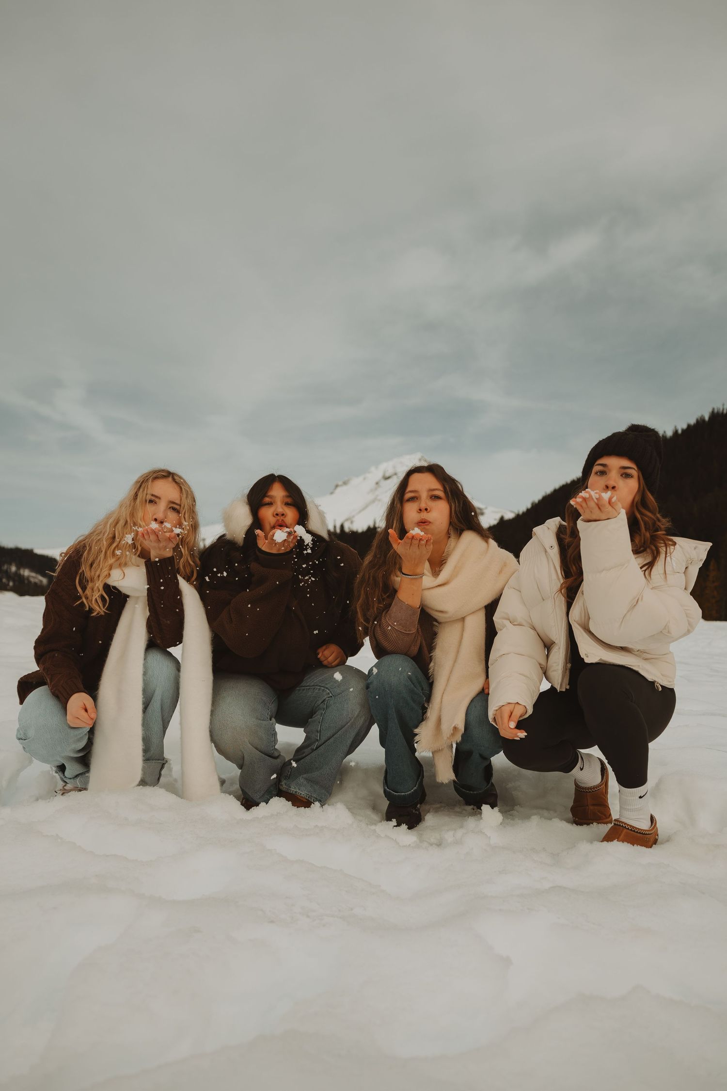 Four women kneel in snow blowing kisses with snowy mountain behind them.