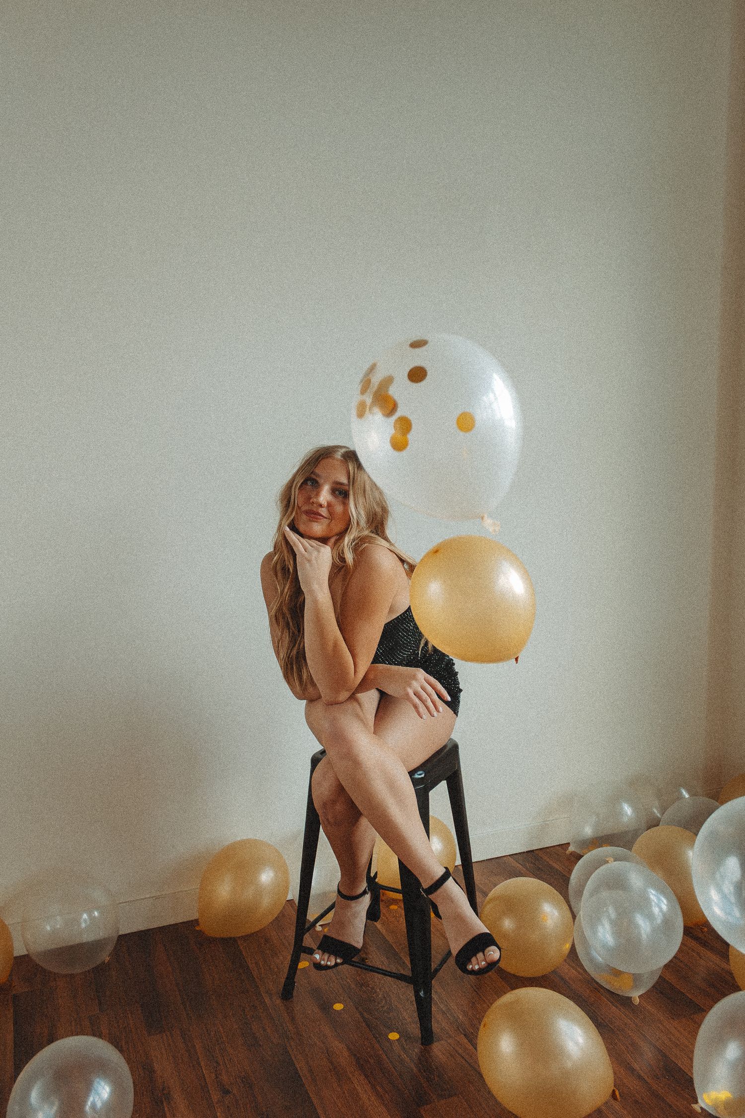 Stylish photoshoot with gold and white balloons featuring someone posing on a wooden stool against neutral wall.