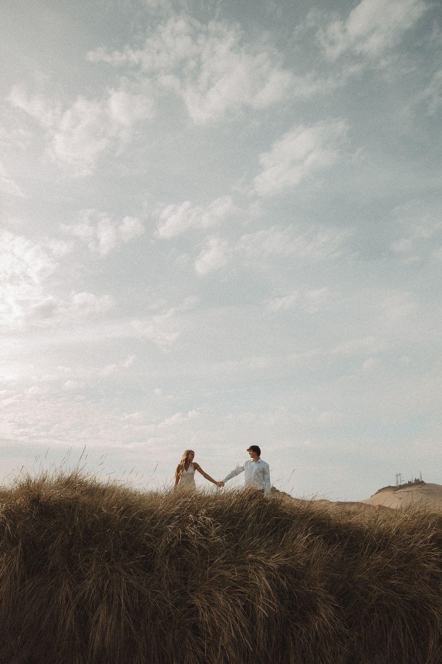 Couple holding hands on sandy dunes with tall grass under a cloudy sky.