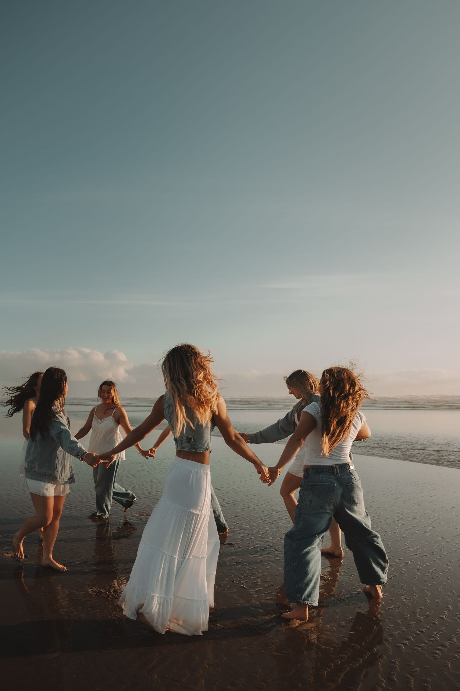 Five women hold hands dancing in a circle on a sunlit beach with ocean backdrop.