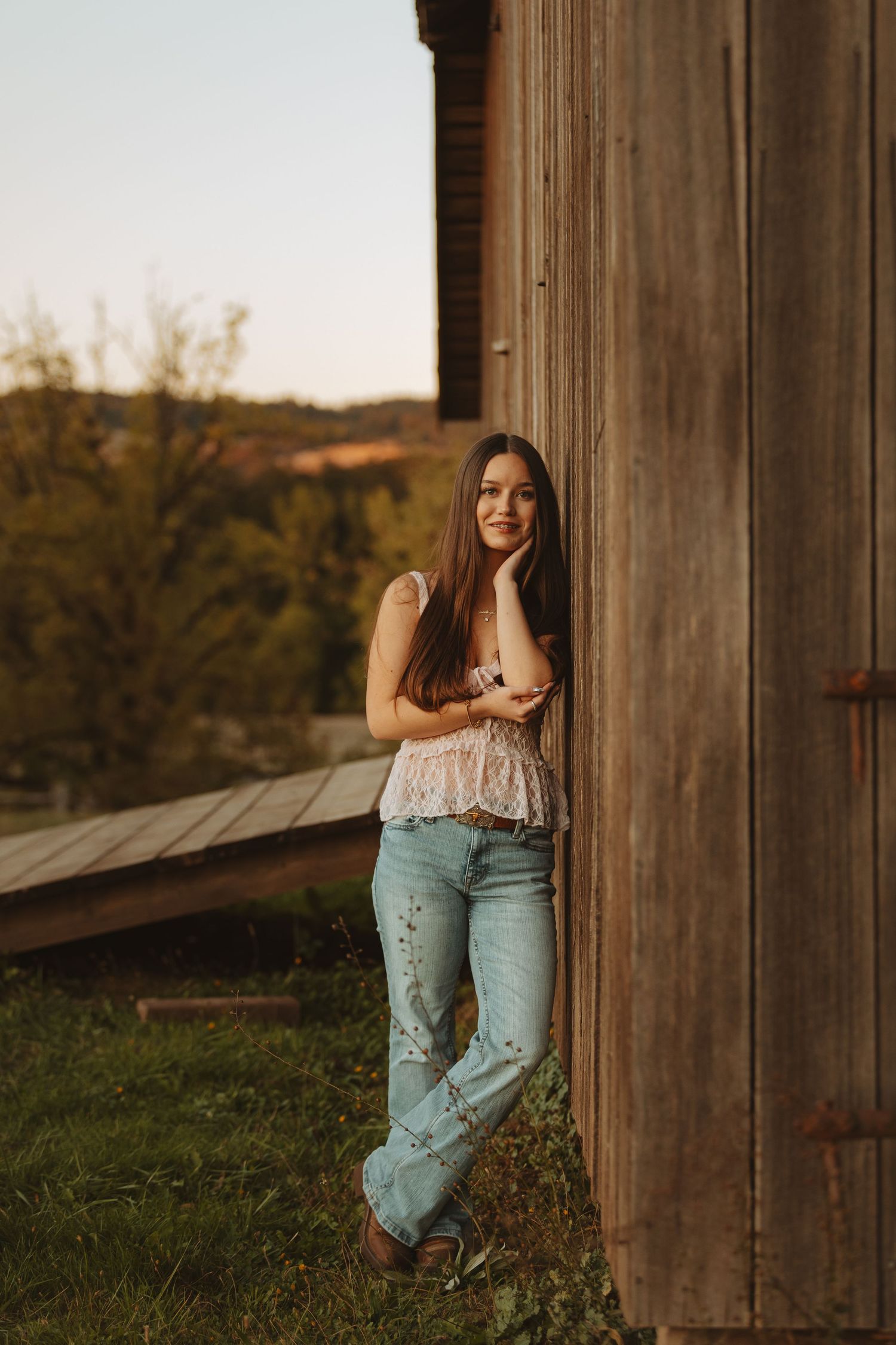 Young woman in lace top and wide-leg jeans leaning against rustic wooden barn at golden hour sunset.