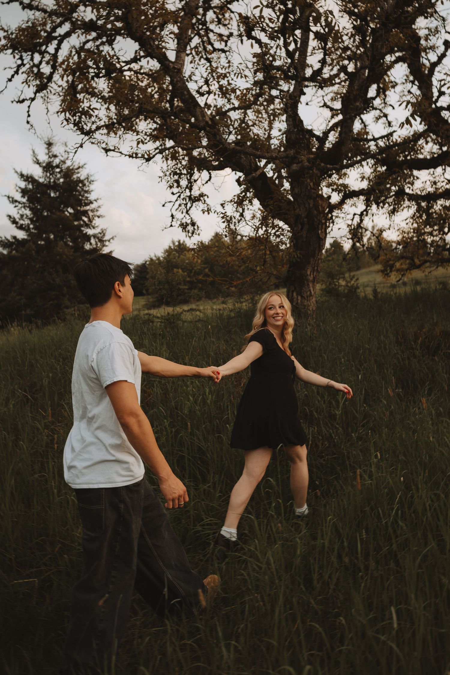 A couple holds hands and dances playfully in a green meadow beneath a large tree at dusk.