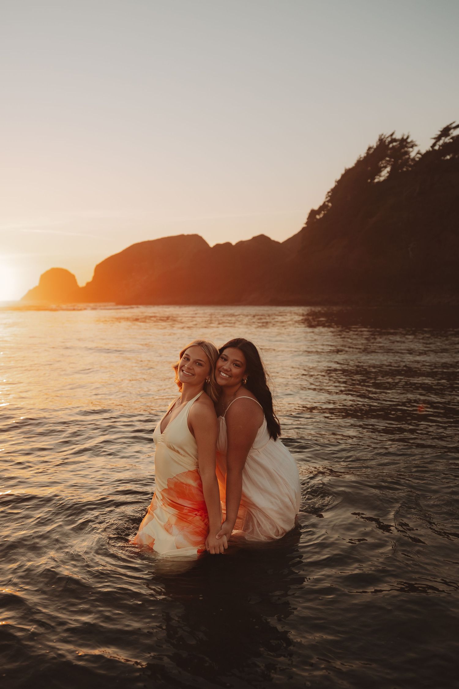 Two women in white dresses stand in ocean water at sunset with rocky cliffs in the background.