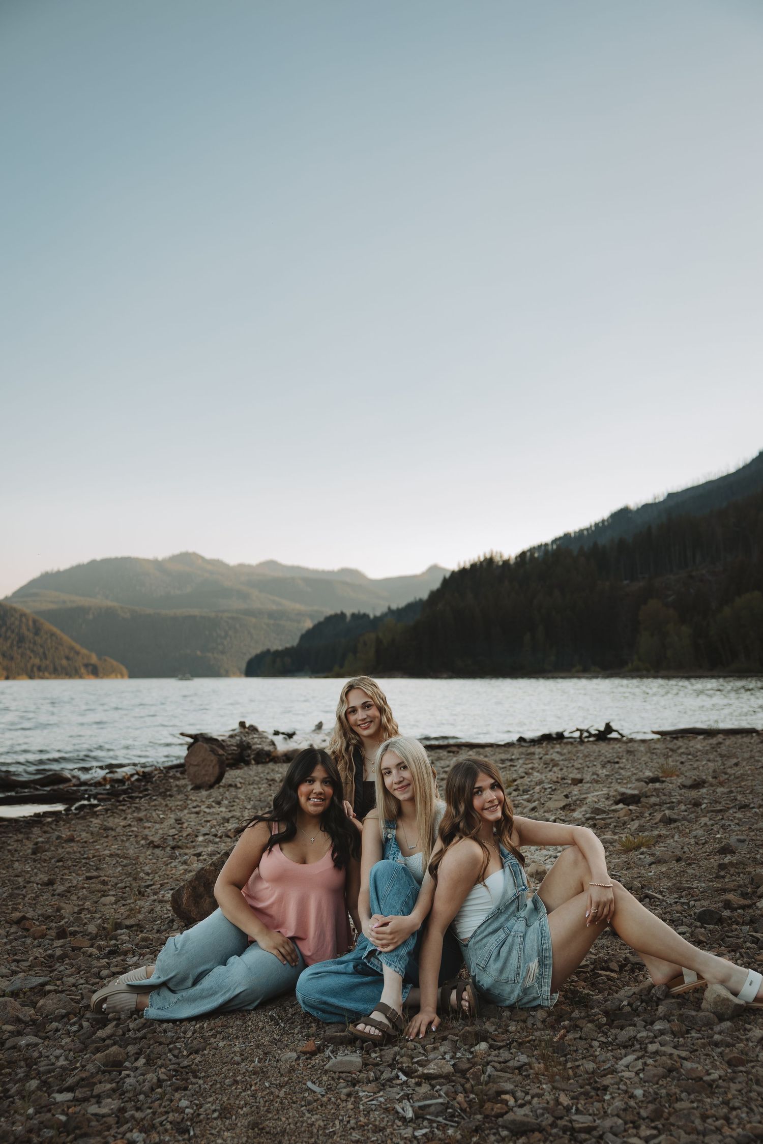 Four young women posing together on rocky lakeside beach with mountain and forest backdrop at dusk.