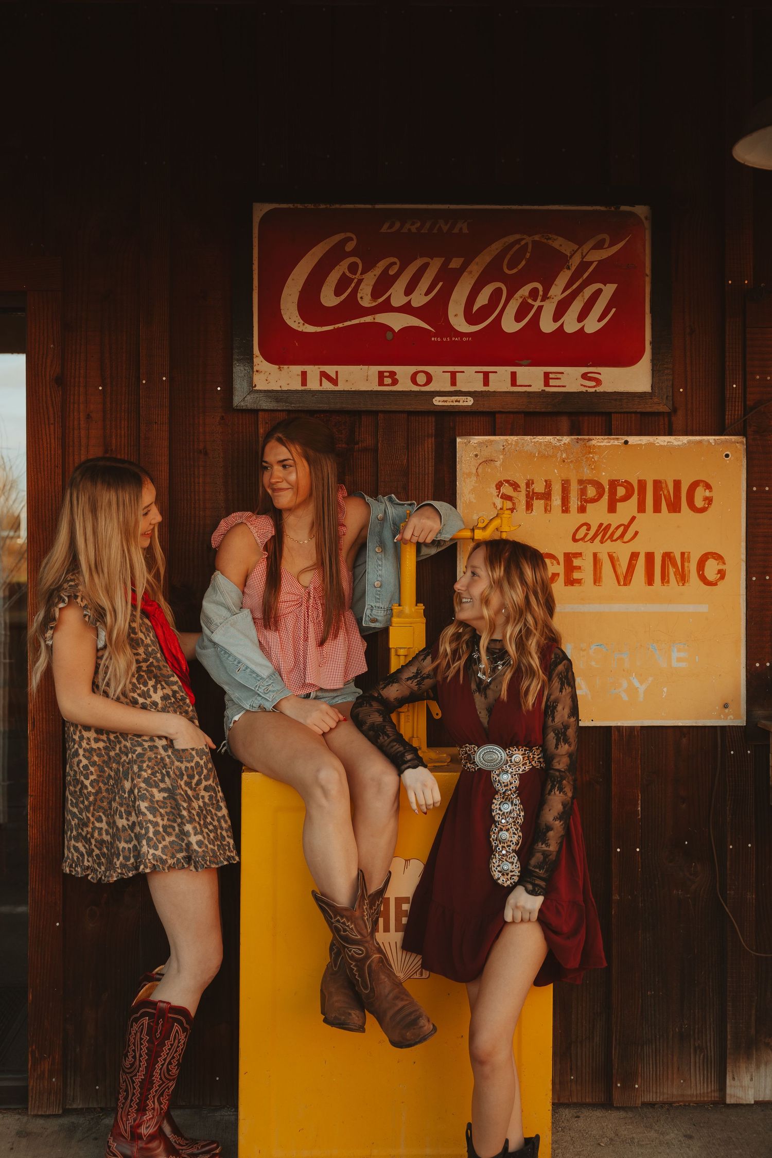 Three young women in western outfits chat near vintage Coca-Cola and Shipping signs at rustic location.
