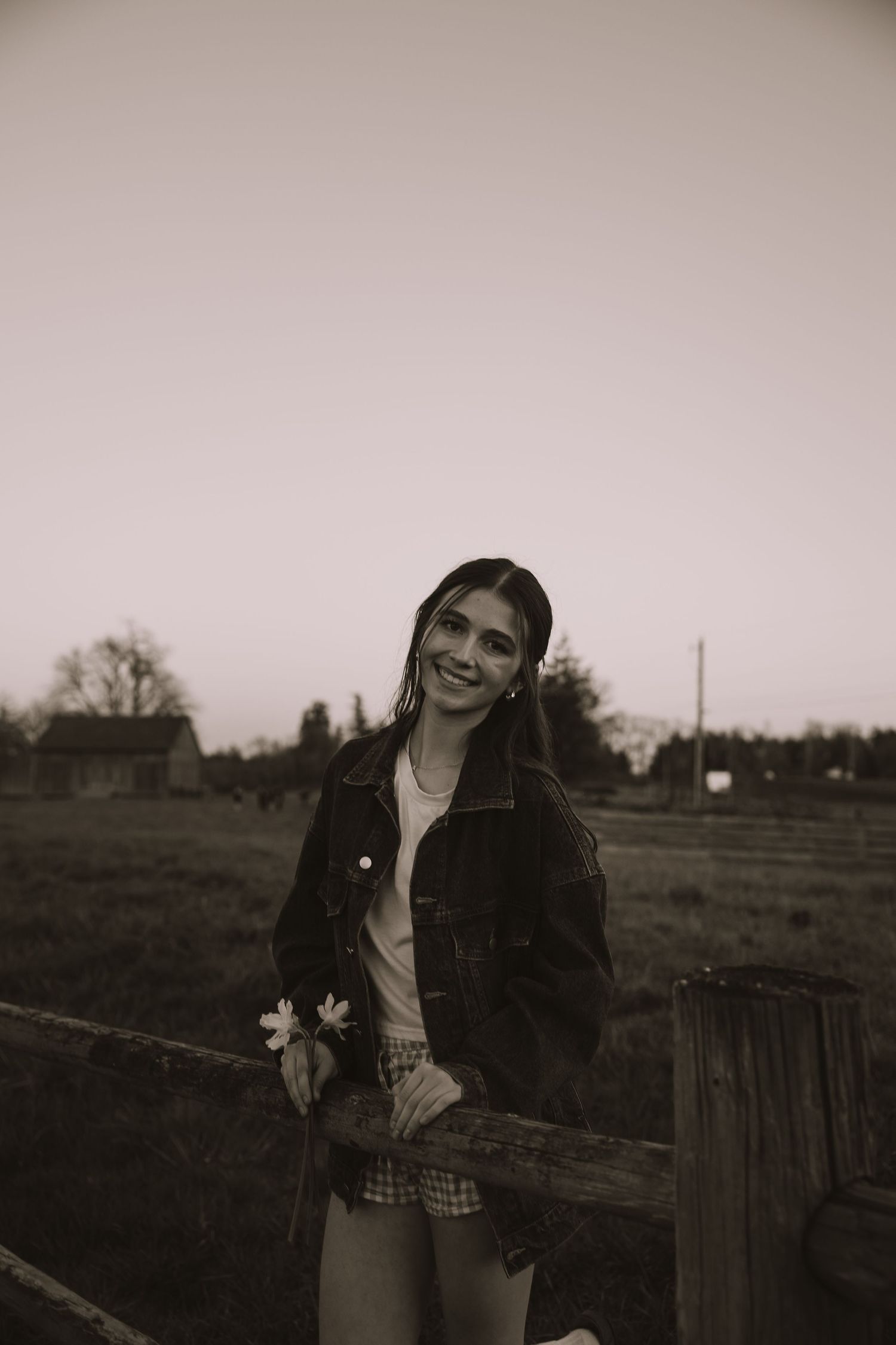 Black and white photo of a smiling young woman in a denim jacket holding flowers by a rustic wooden fence in a rural field.