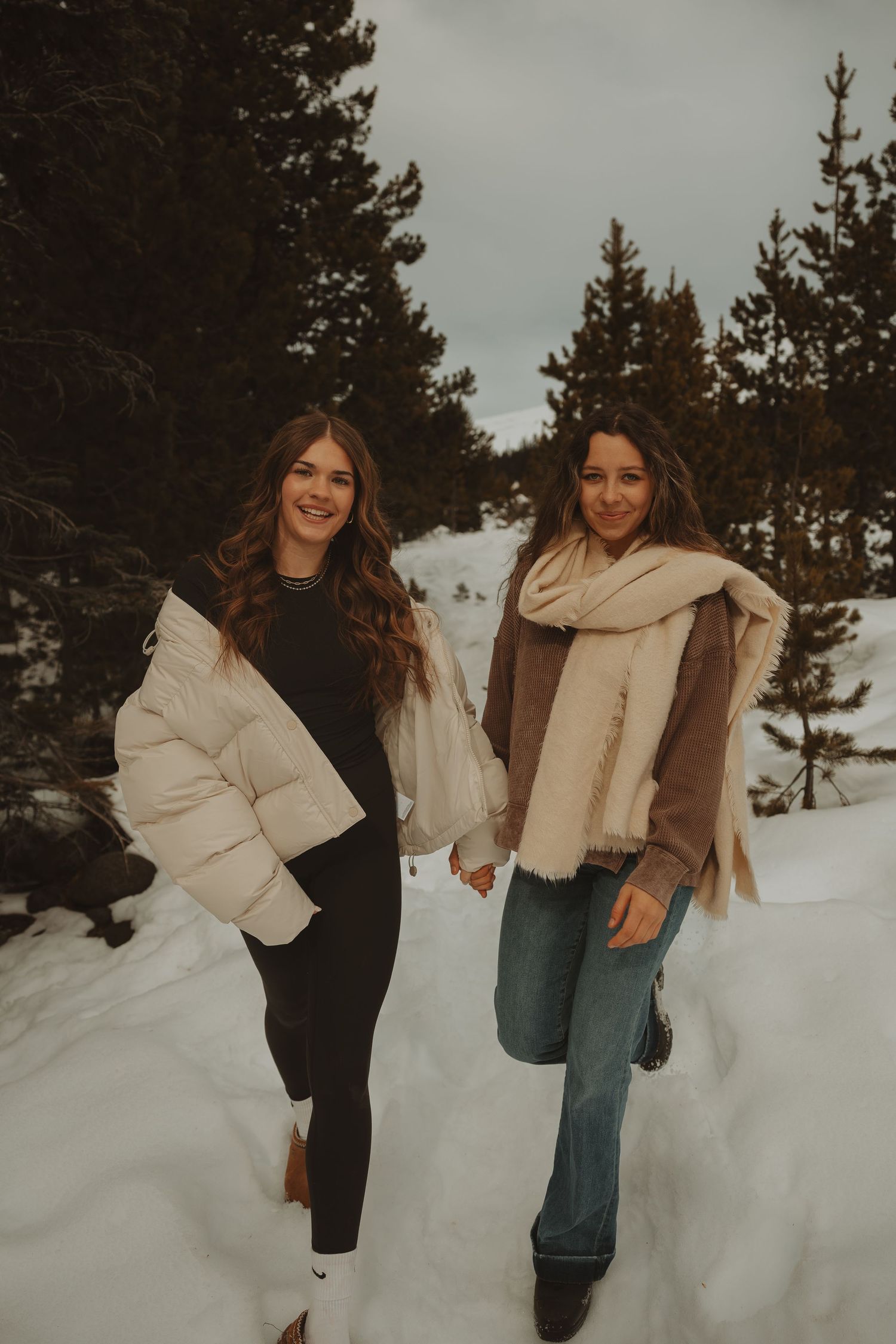 Two smiling women hold hands walking through snowy pine forest in winter coats and scarves.