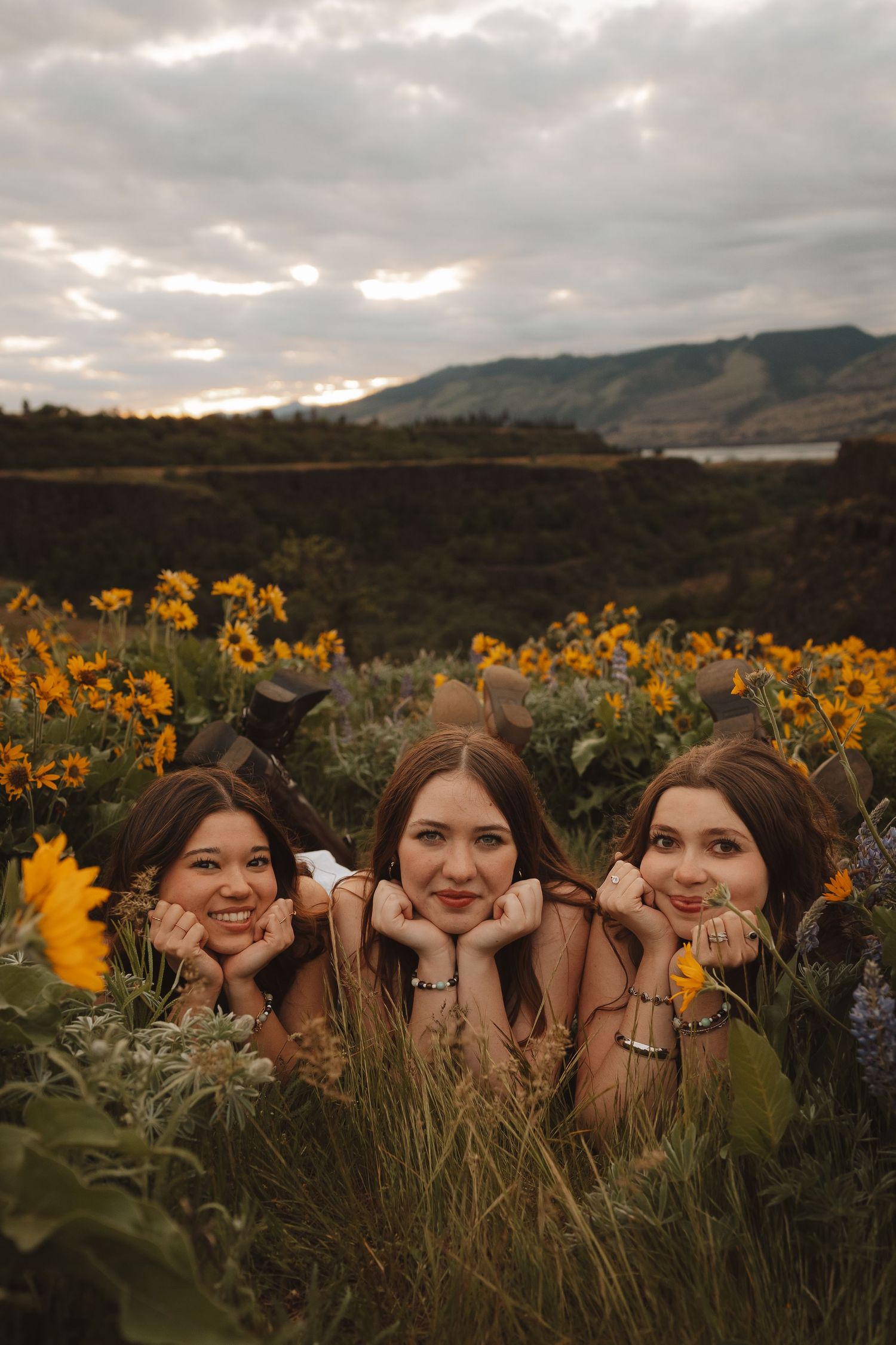 Three young women lying in a sunflower field with mountains and cloudy sky in the background.
