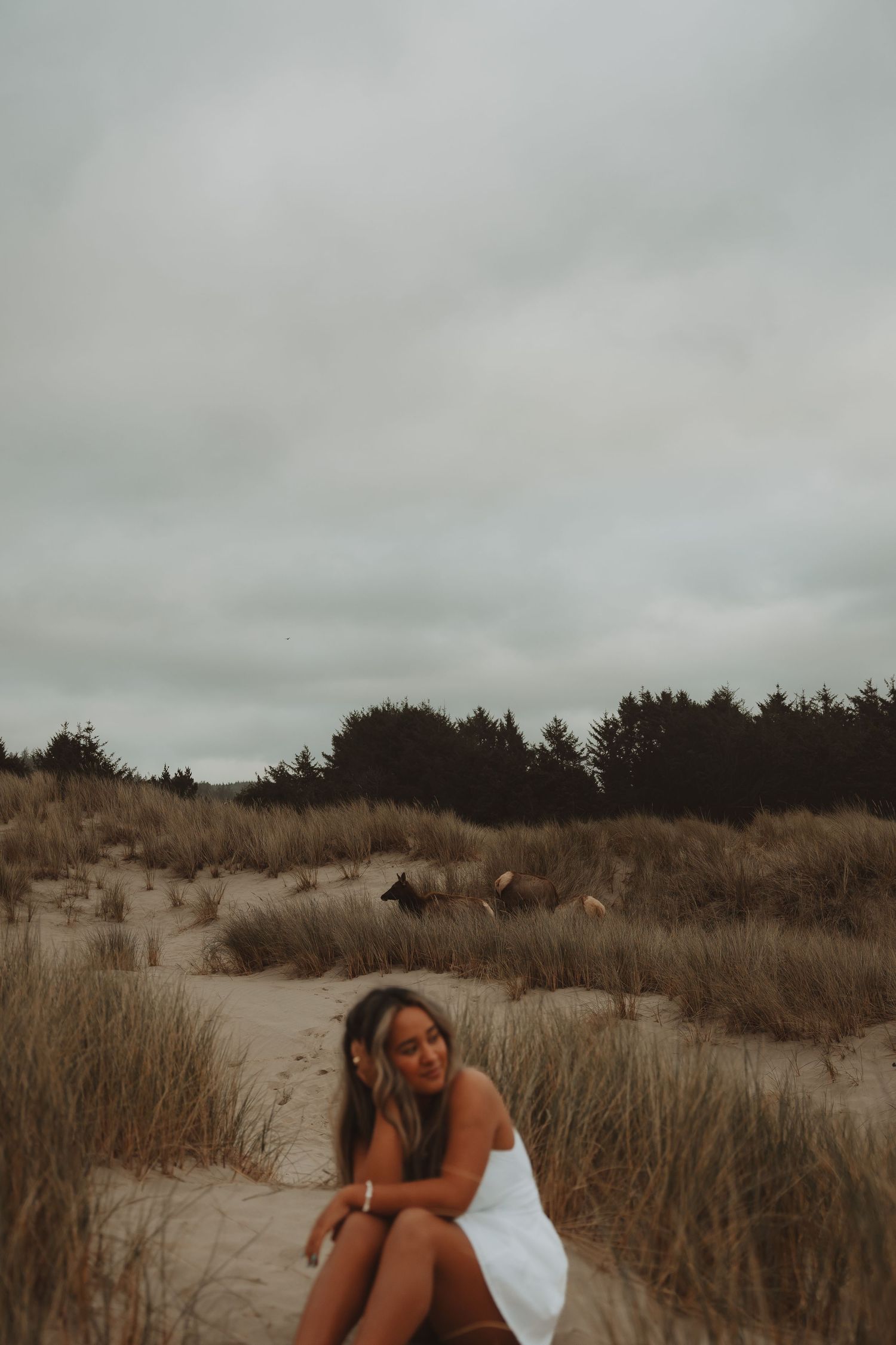 Woman in white dress sitting on sand dunes with trees and cloudy sky in the background.
