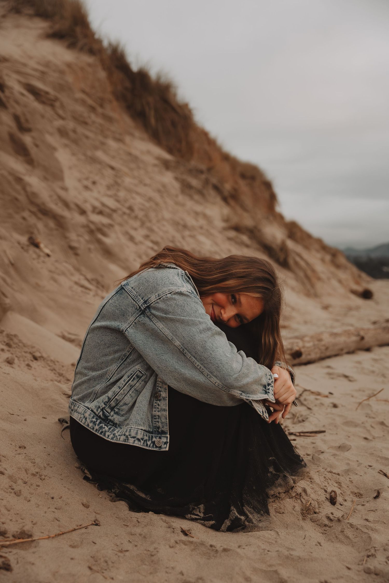 Person in denim jacket lies artistically on sandy beach dunes during cloudy weather.