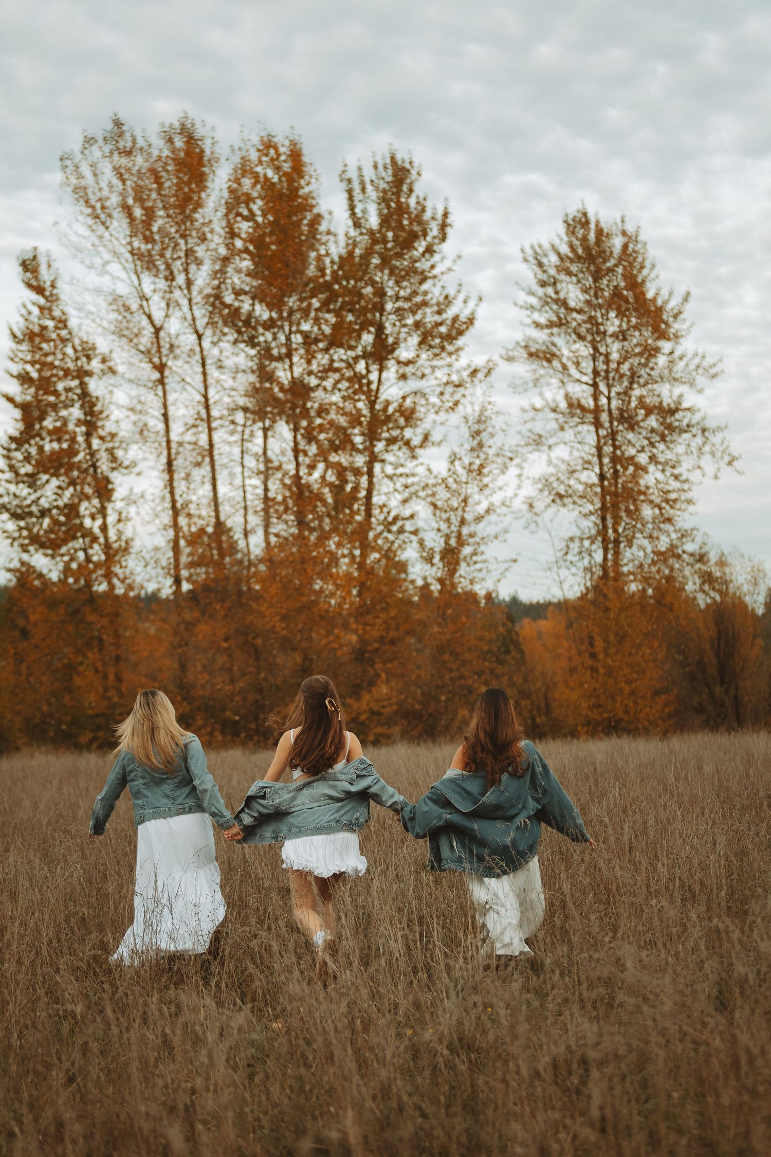 Friends in denim jackets and white dresses walk through a golden autumn field with tall trees in background.