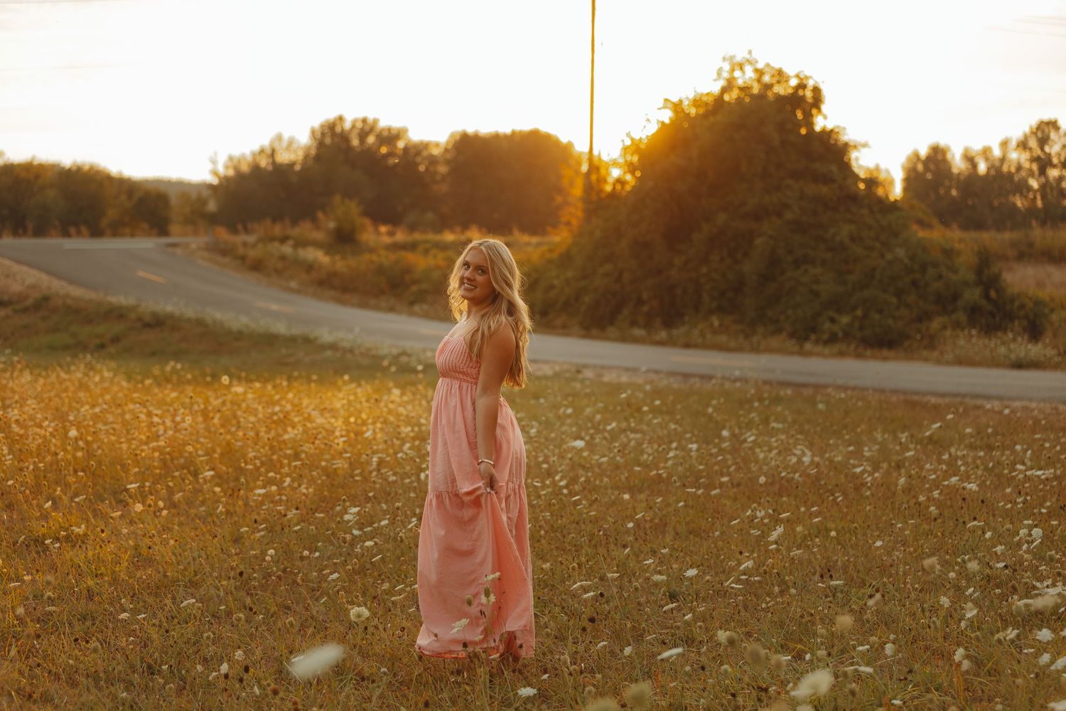 Woman in pink maxi dress standing in golden wildflower field at sunset with warm glowing light.