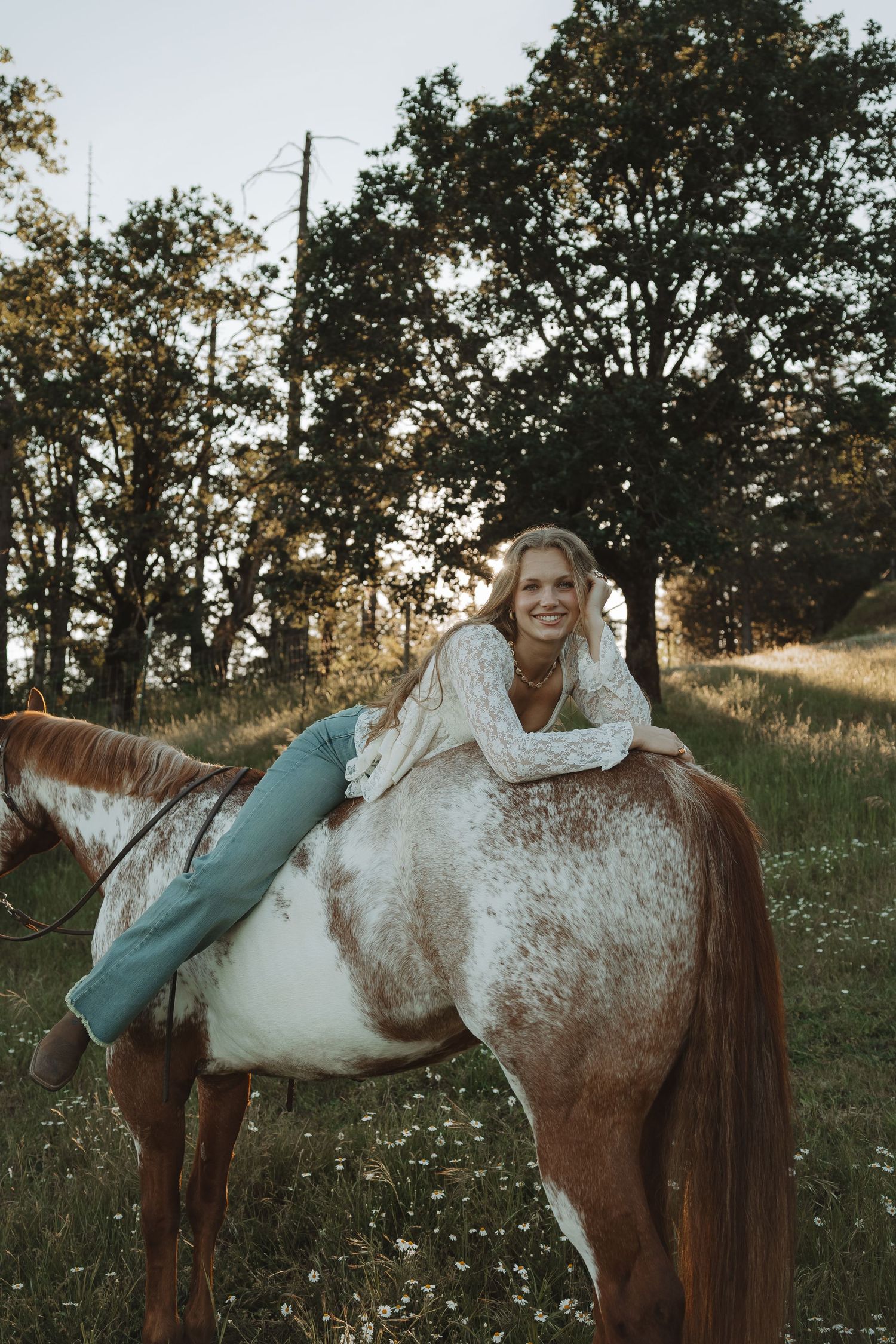 Smiling blonde woman lying on a brown and white horse in a scenic green meadow with trees at golden hour.