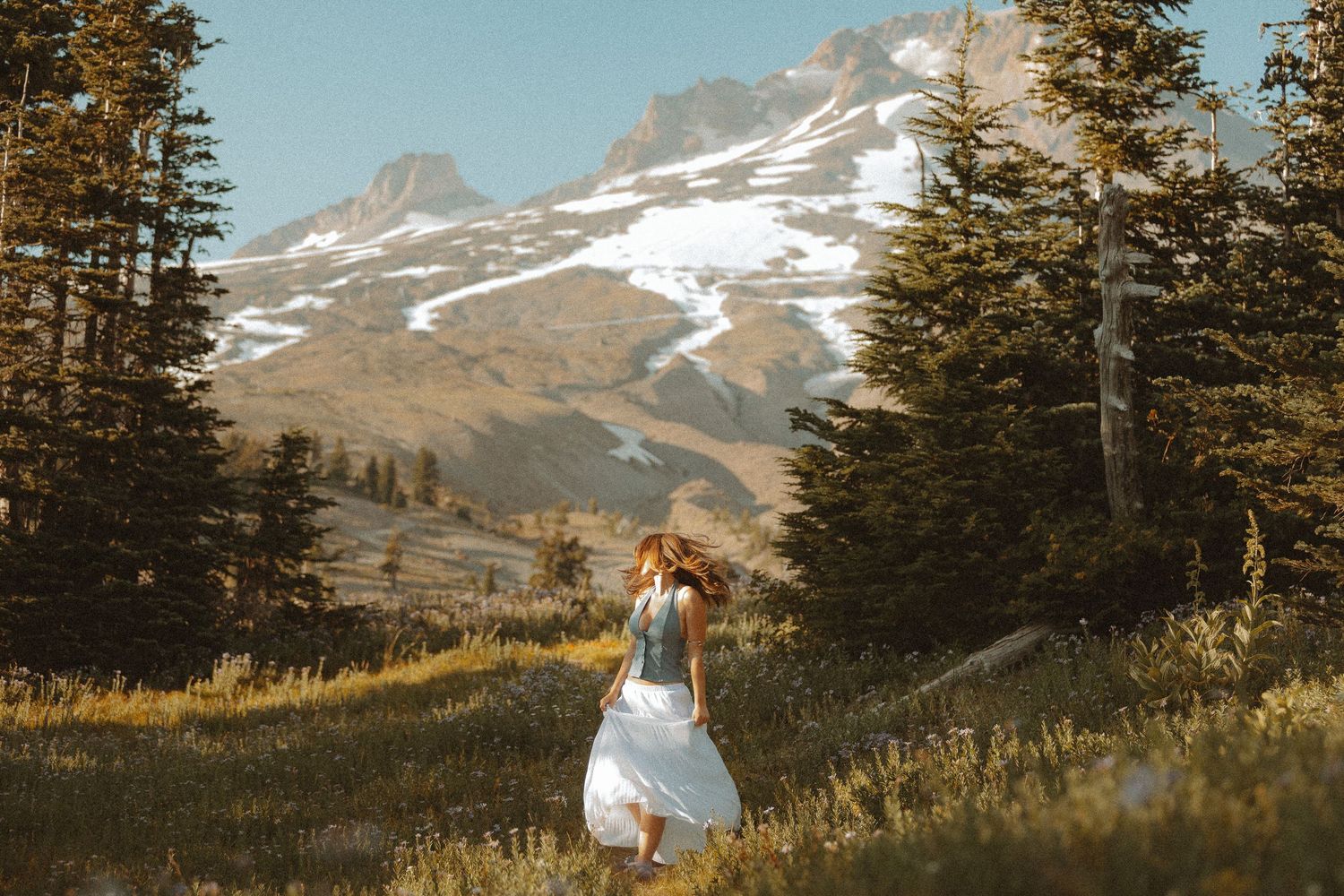 Woman in white dress walking through alpine meadow with snow-capped mountain and evergreen trees in background.