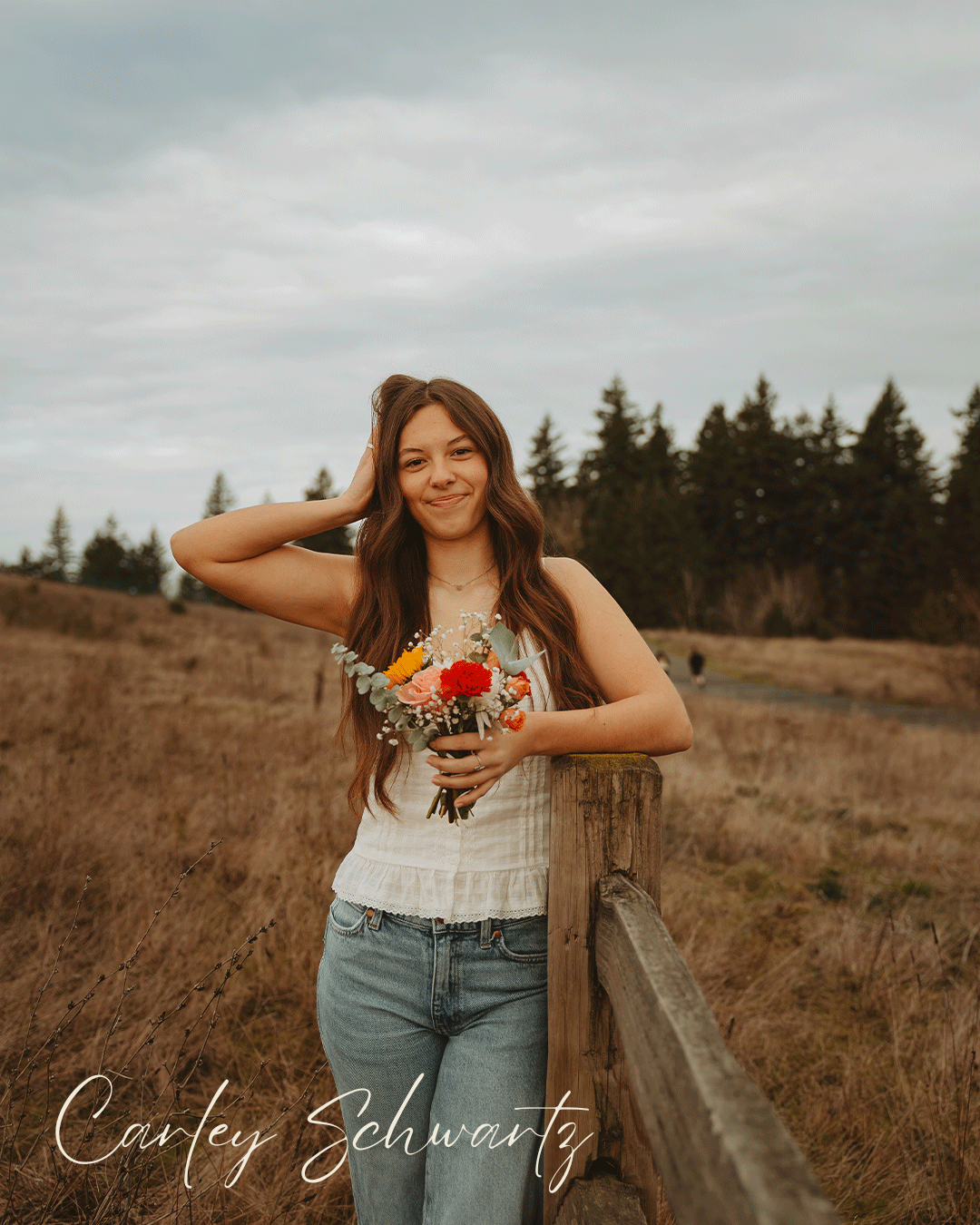 A person in casual attire poses in a rustic outdoor setting with autumn-toned fields and evergreen trees.