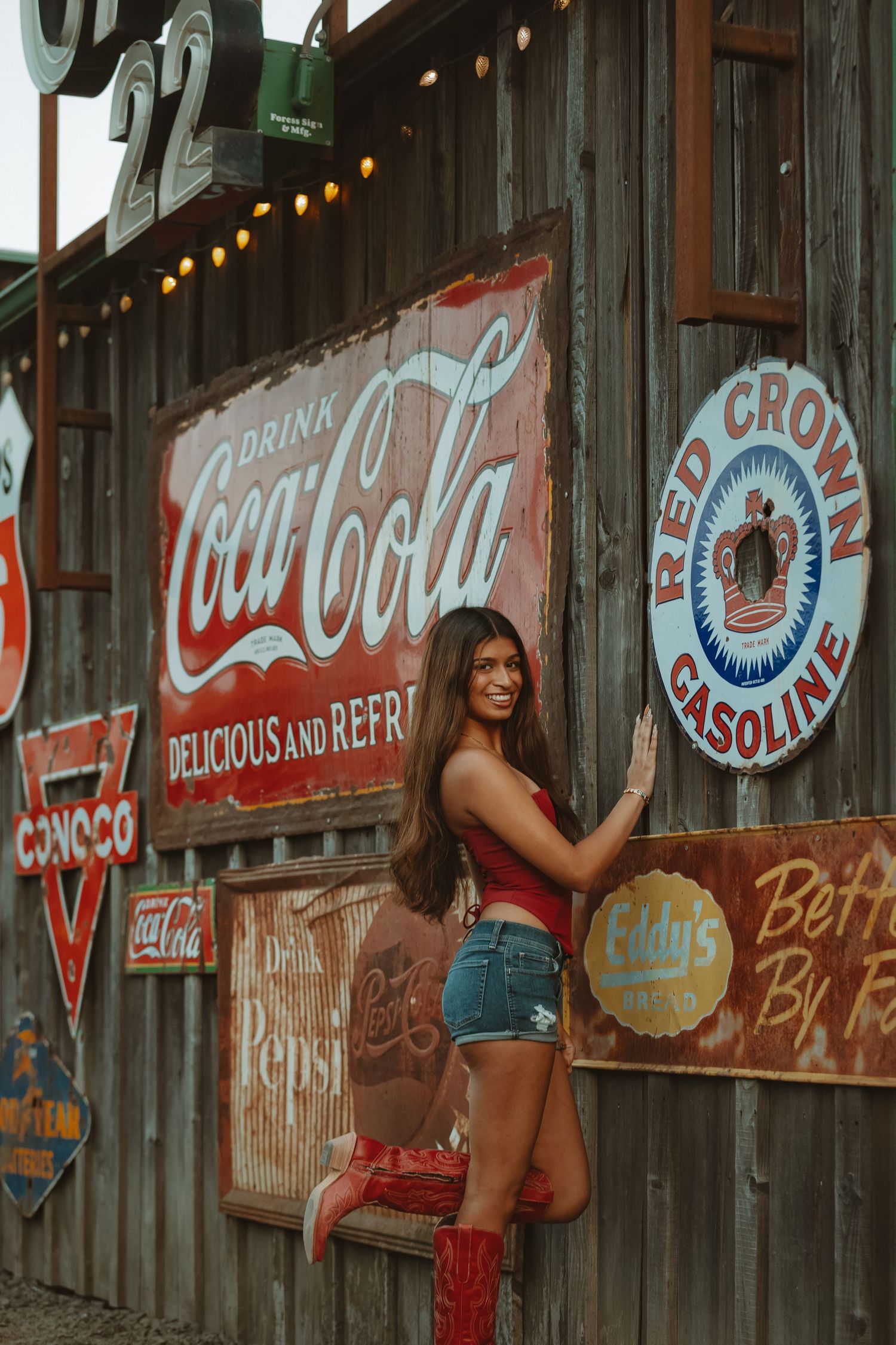Collection of antique advertising signs on weathered wooden wall with string lights.
