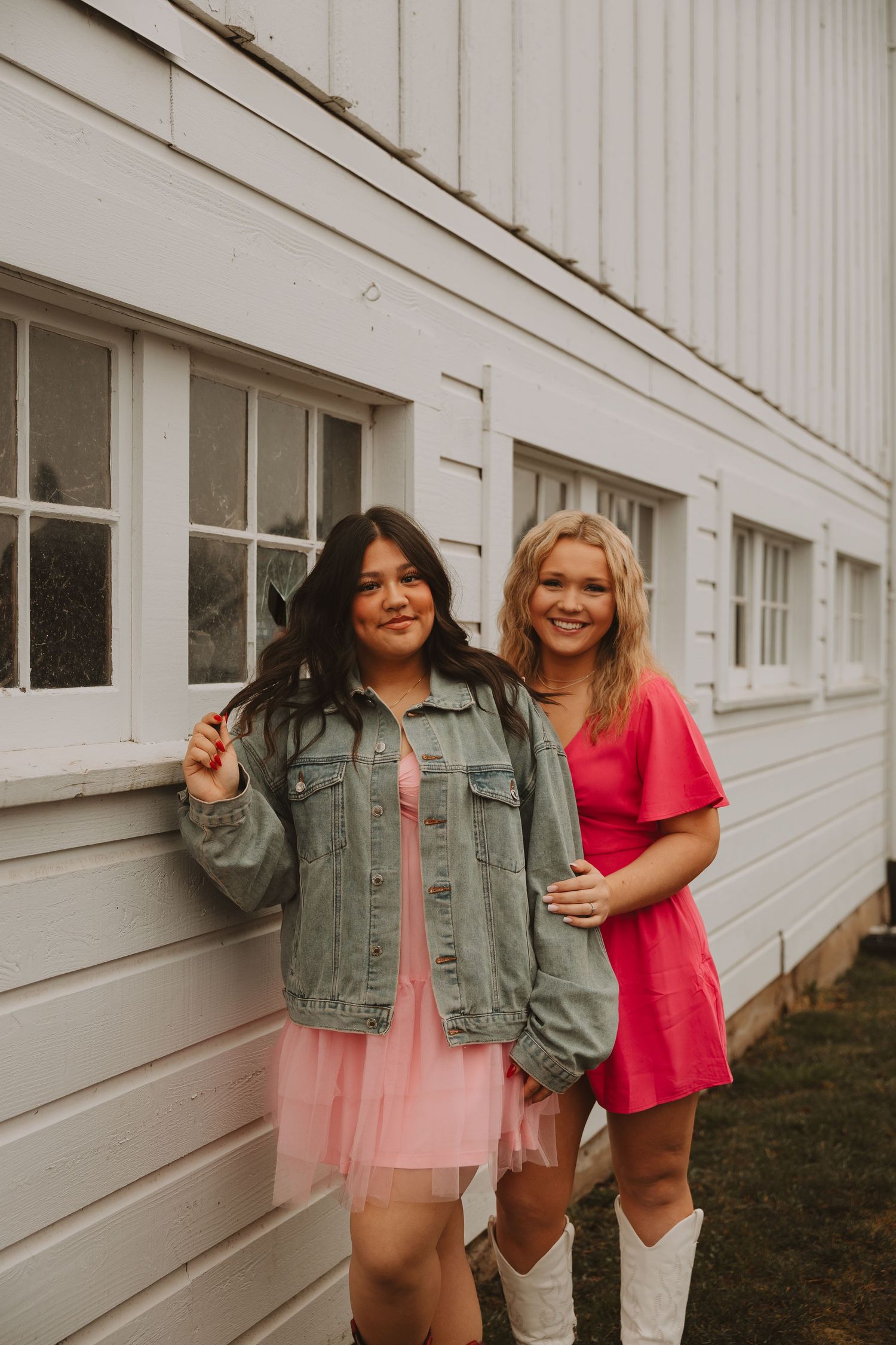 Two friends in casual outfits laughing and posing playfully against white building wall.