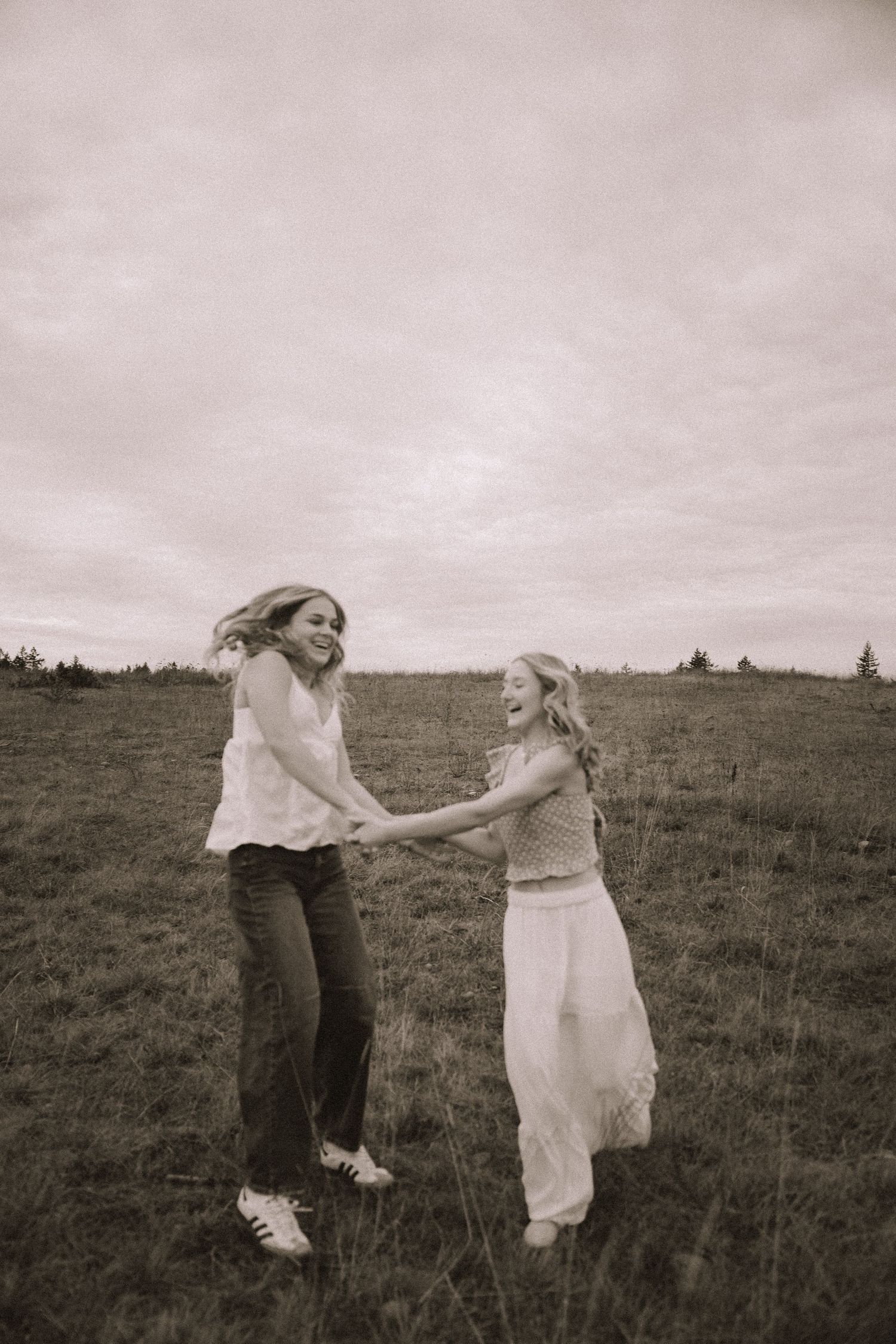 Two people playfully dance together in a grassy field under moody skies in black and white.