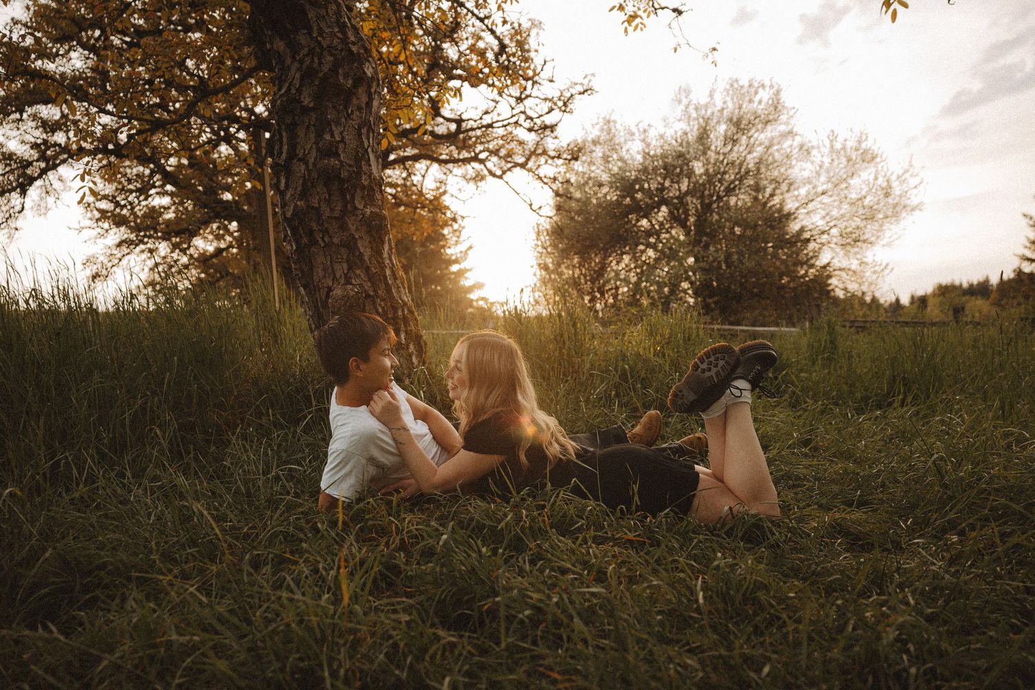 Couple sitting together in golden hour sunlight by a tree in a lush green meadow, sharing a romantic moment.