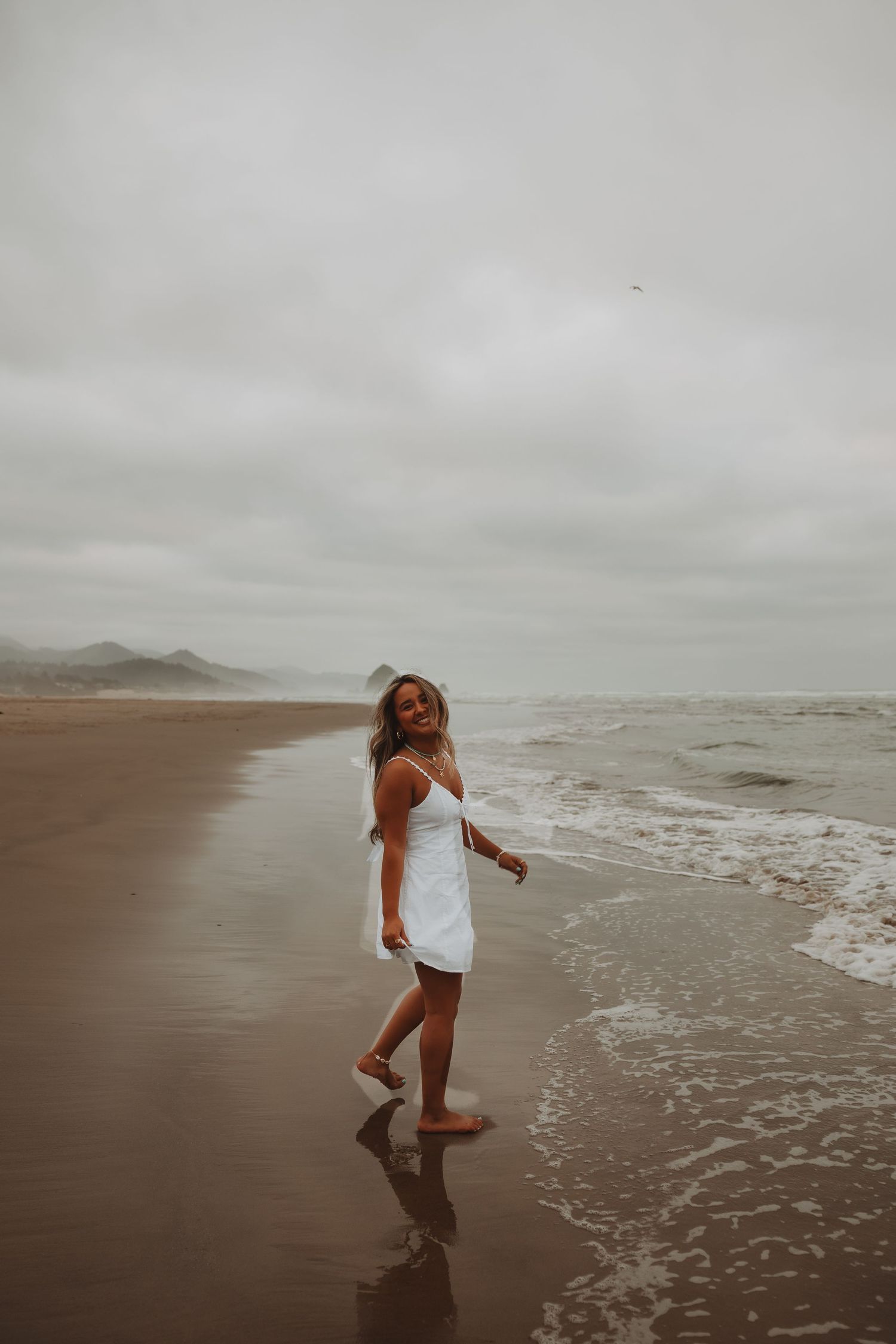 Woman in white sundress walking barefoot along sandy beach shore on a cloudy day.
