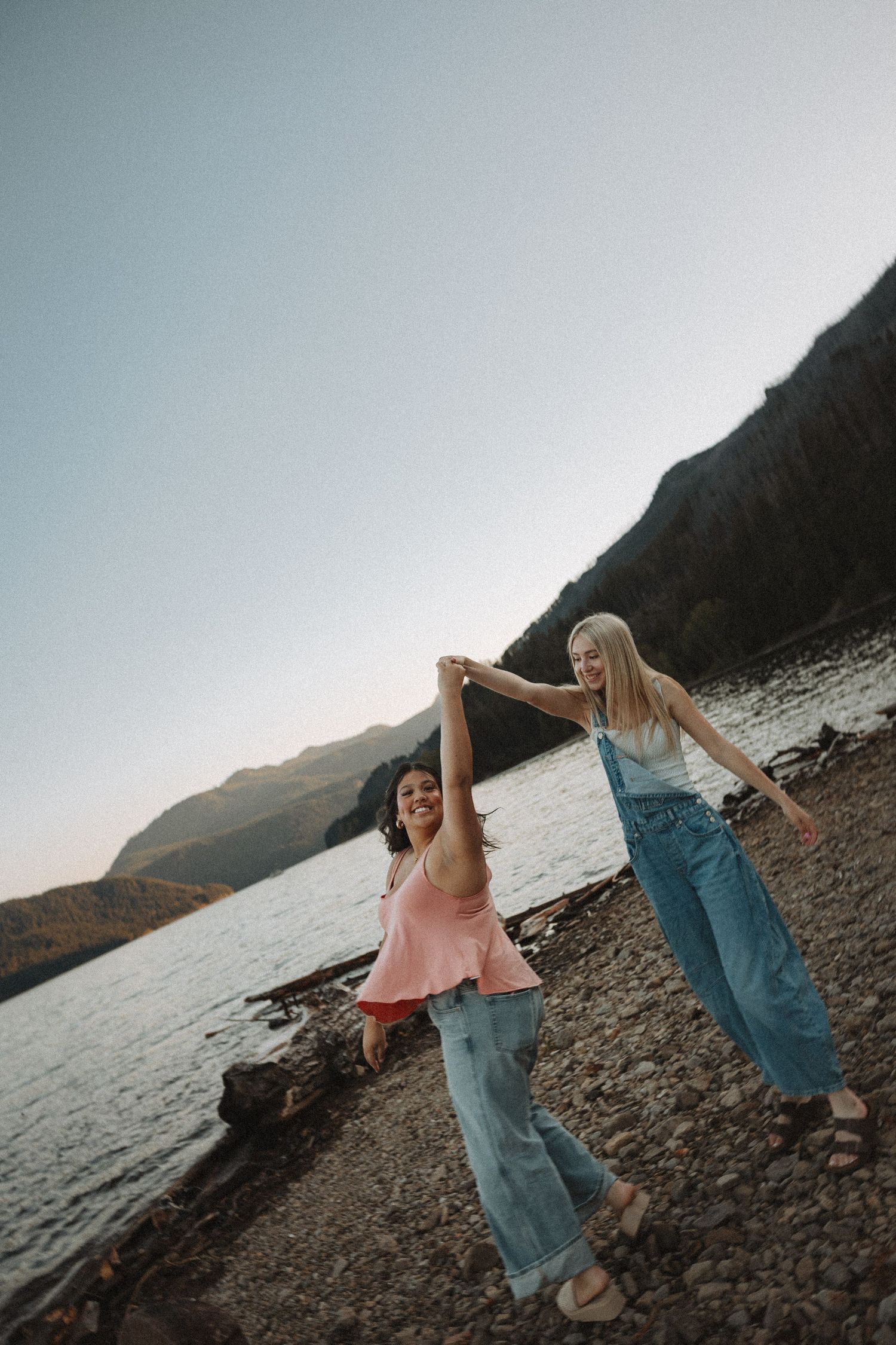 Two young women holding hands and dancing on a rocky mountain lakeside at dusk.