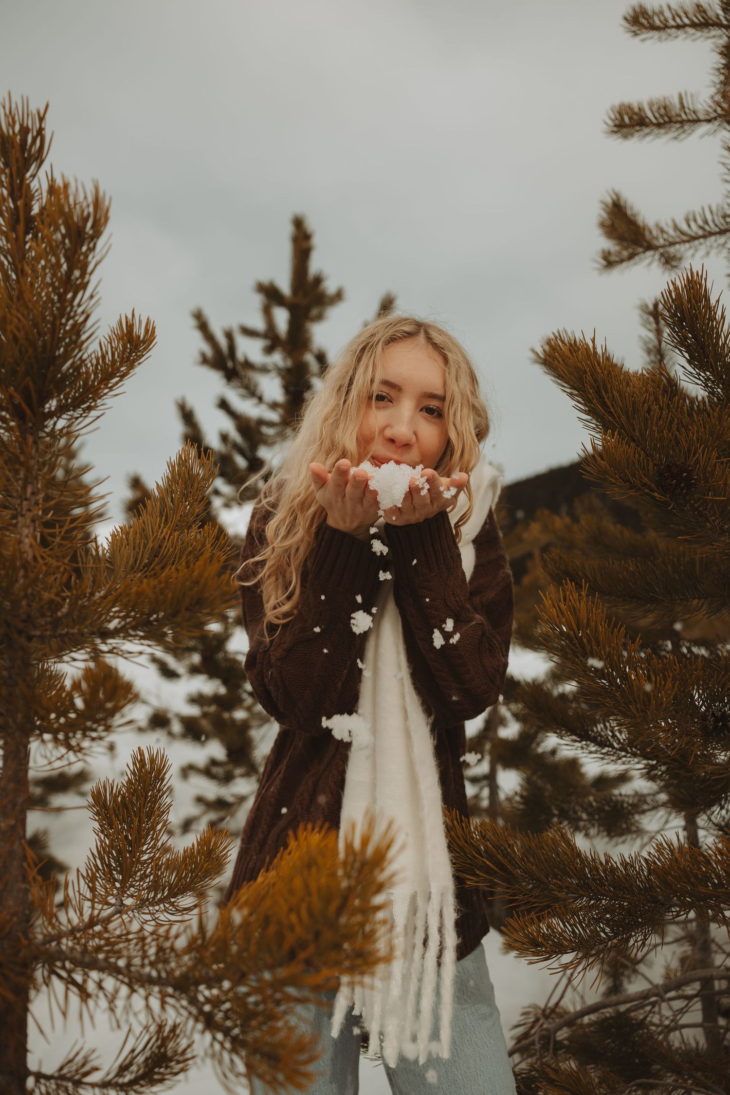 Blonde woman blowing snow from her hands outdoors among pine trees in winter, wearing a brown coat and white scarf.