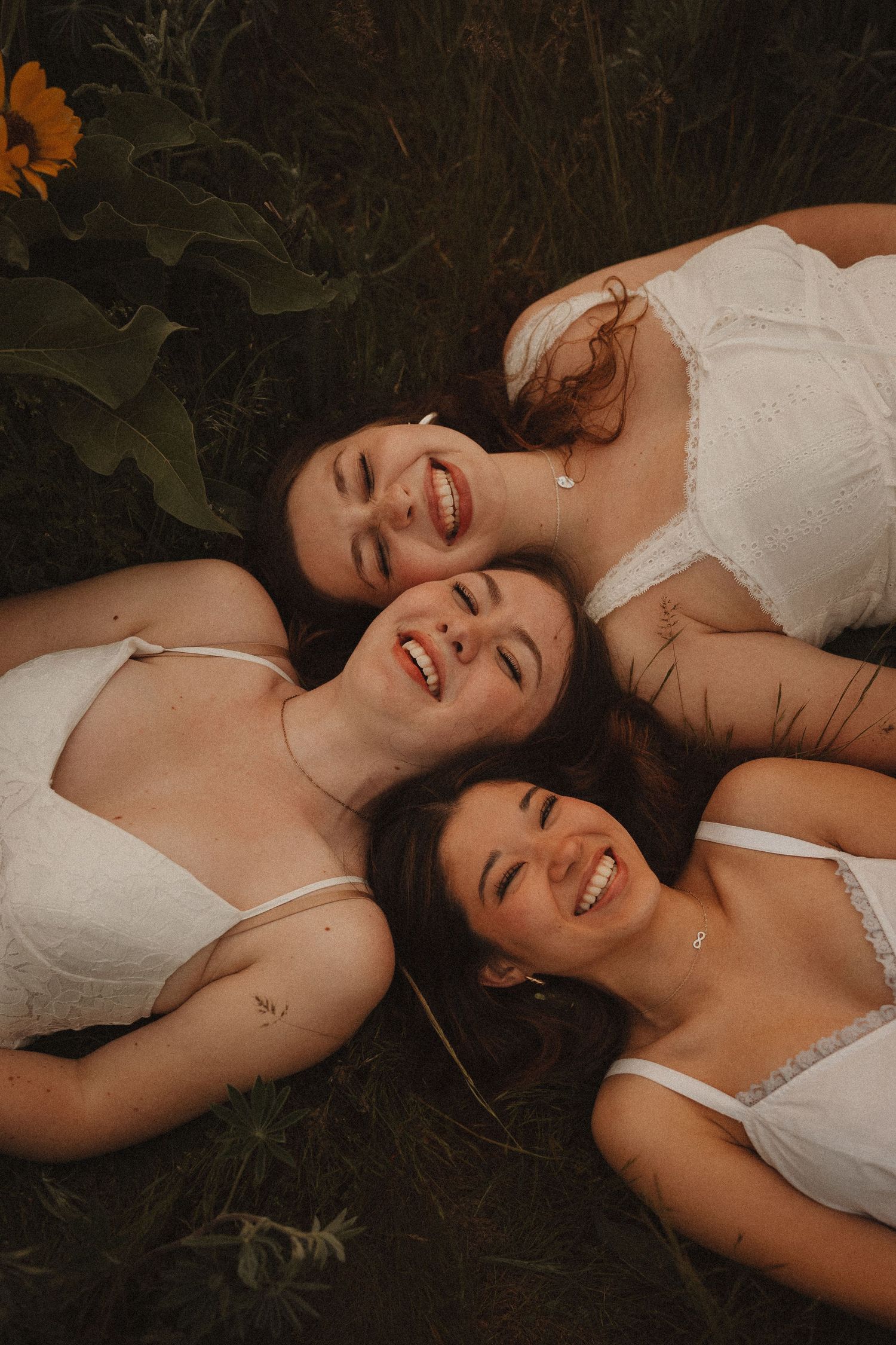 Three smiling women lying in grass wearing white tops with sunflowers blooming nearby.