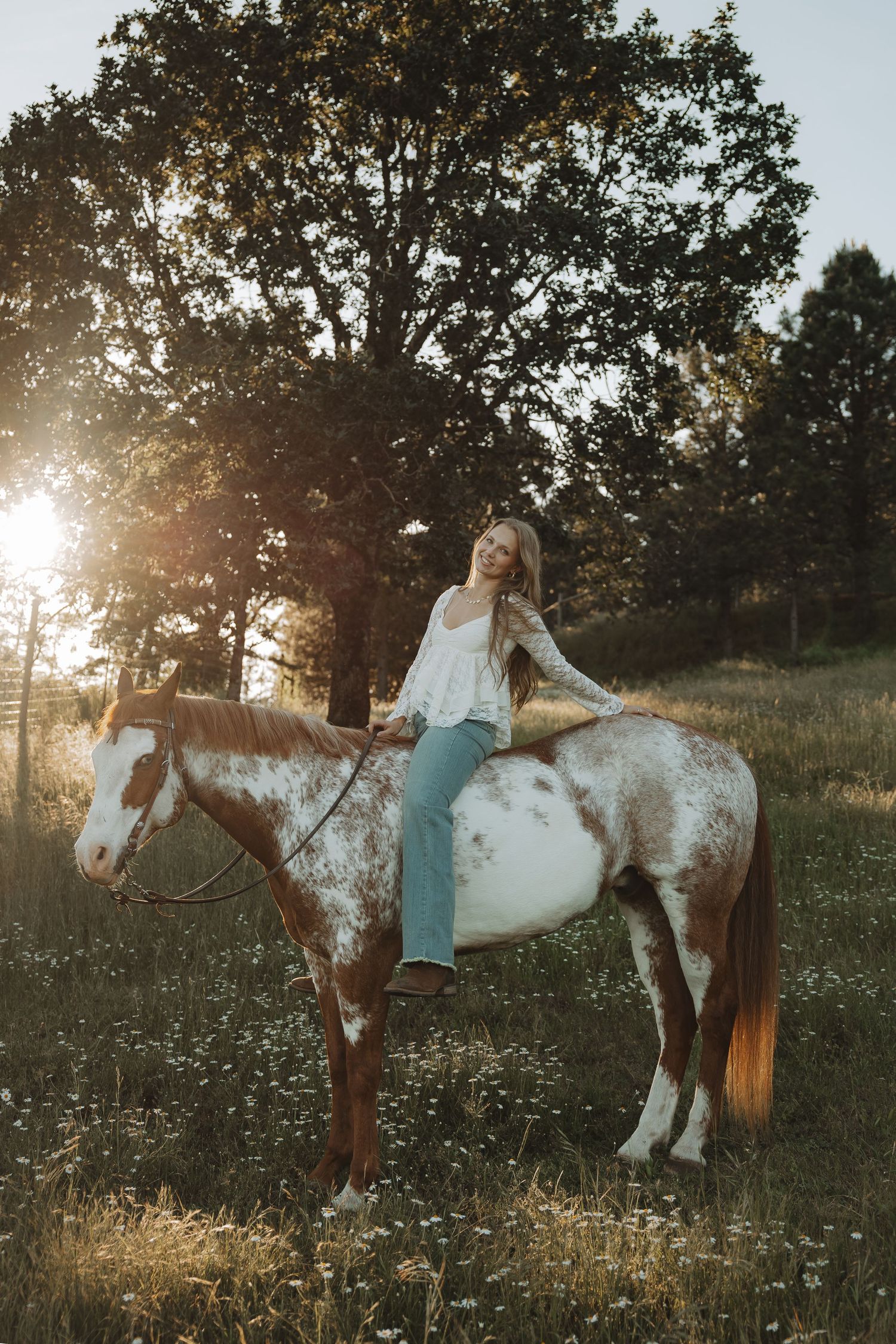 Young woman in white top and jeans riding a brown and white paint horse in a sunlit meadow.