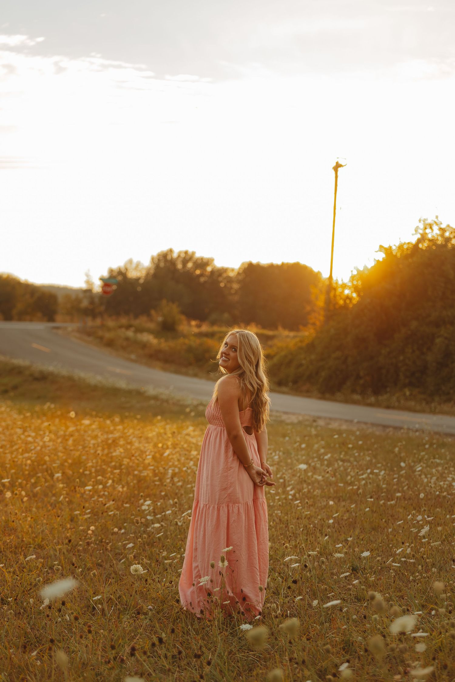 Woman in pink maxi dress standing in golden hour sunlit field with wildflowers and road in background.