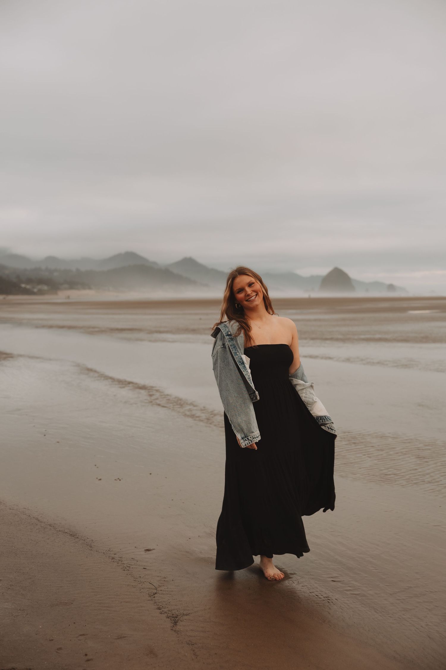 A figure in a flowing black dress twirls gracefully on a misty beach with mountains in the background.