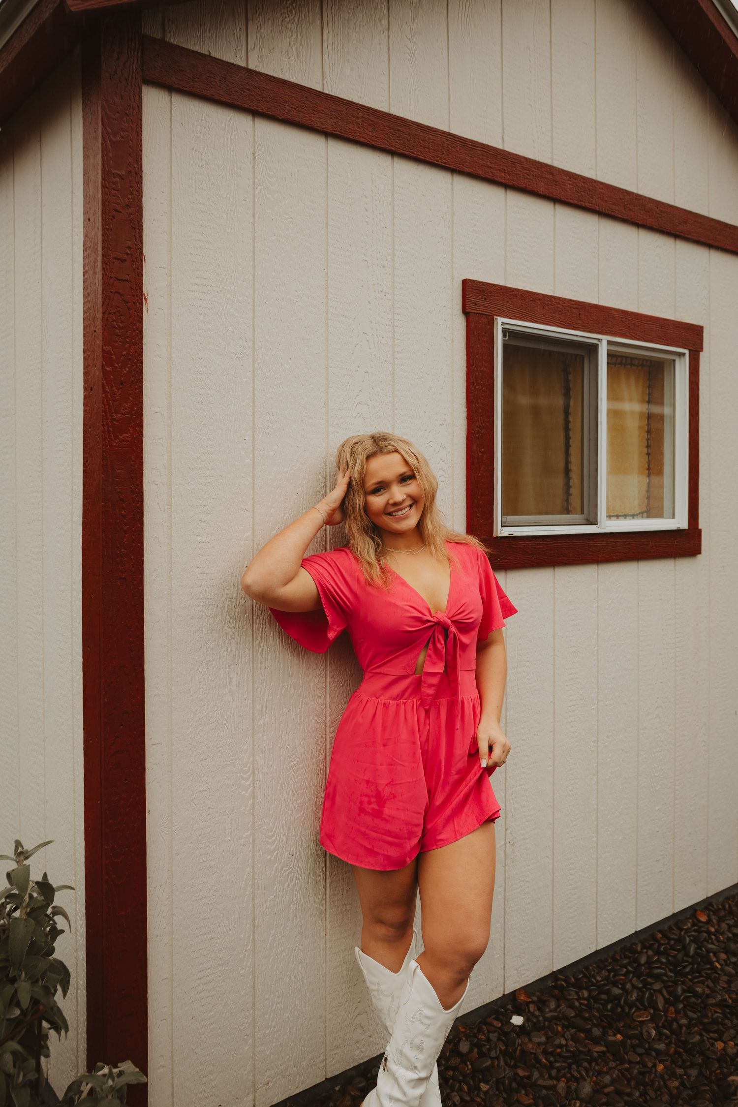 Person wearing red romper and white boots poses casually against beige building wall.