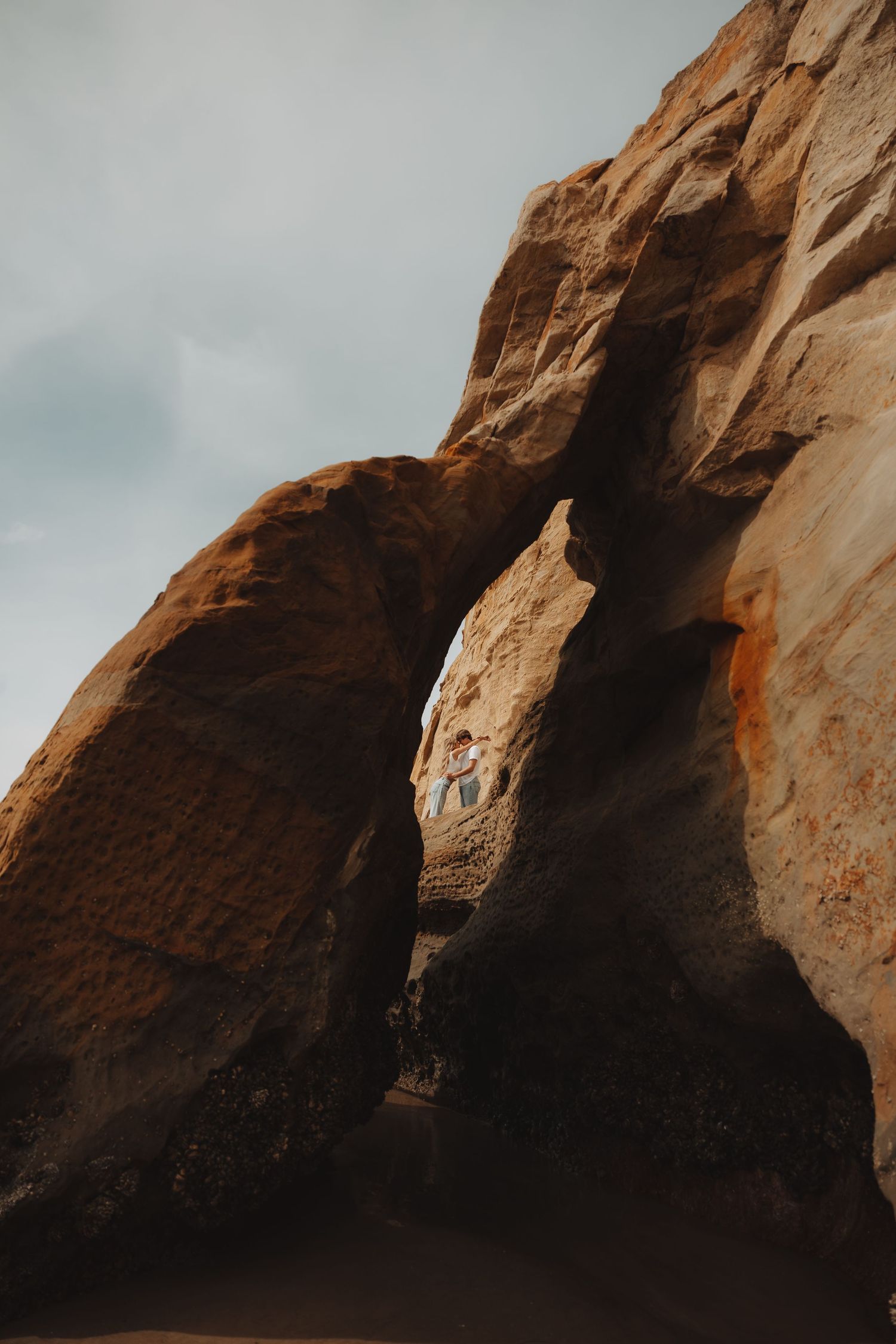 Couple stands framed within a dramatic natural sandstone rock arch formation under cloudy skies.