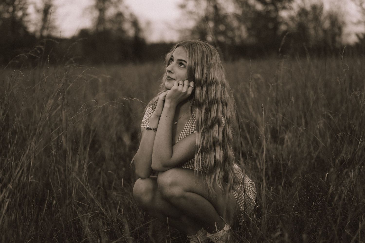 Someone sits contemplatively in tall grass during golden hour with trees in the distance.