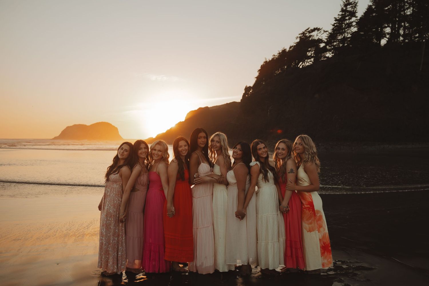Group of bridesmaids in colorful dresses posing on a beach at golden sunset with ocean waves and rocky cliffs.