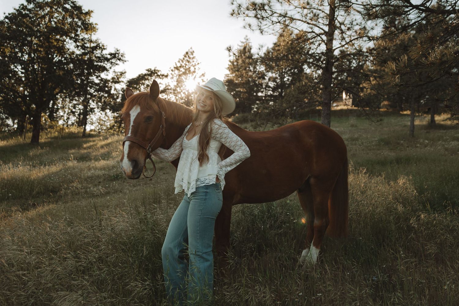 Woman in cowboy hat and jeans stands beside a chestnut horse in a sunlit meadow surrounded by trees.