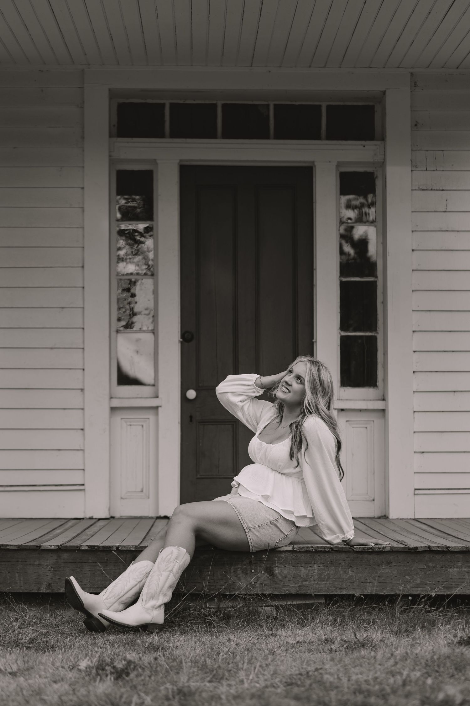 Blonde woman in white outfit and cowboy boots sitting on porch steps in black and white photo.