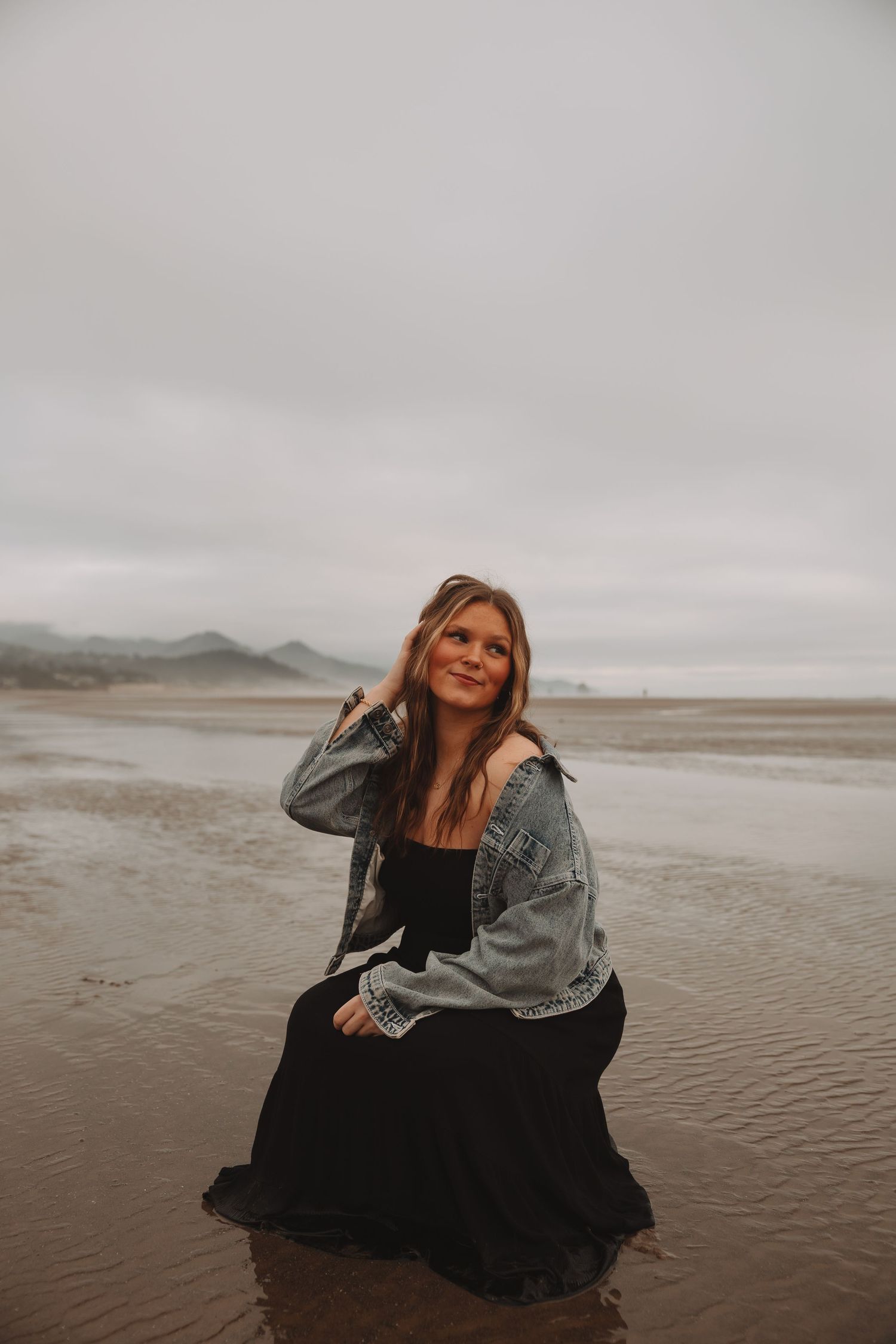 Person in denim jacket and black dress sits on wet sand at a misty coastal beach during cloudy weather.
